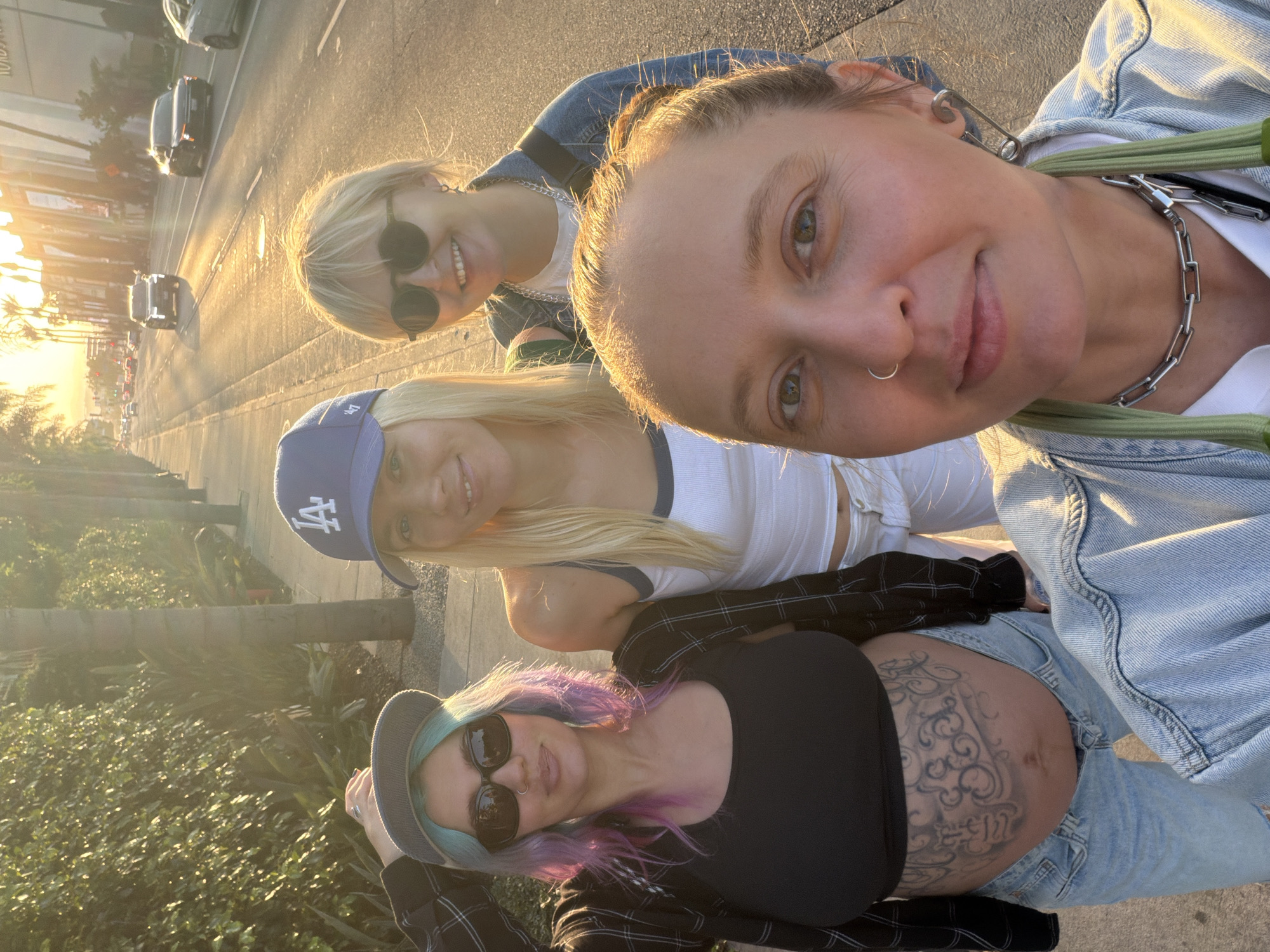 Four women are photographed at street level under bright sunlight, which casts shadows across the frame. The camera is positioned at a high angle, suggesting a selfie perspective.
The woman in the foreground, closest to the camera, is wearing a denim jacket over a white top. She has her hair up and is adorned with a safety pin earring and a silver chain necklace. To her left is a woman with blond hair and sunglasses, wearing a patterned top. Below her is another woman with blond hair, wearing a blue Los Angeles Dodgers baseball cap and a white tank top. At the bottom of the frame, a woman with aqua and purple hair, wearing a hat and sunglasses, is prominently displaying a pregnant belly featuring a tattoo. She is wearing a black top and a flannel shirt.
The background reveals an urban setting. Palm trees and other greenery line the street. In the distance, buildings are visible, with one appearing to have a "RODEO" sign, which suggests a location in Beverly Hills, Los Angeles, California. Cars are seen driving on the road, and the overall scene conveys an outdoor setting in an affluent urban environment during daylight.
