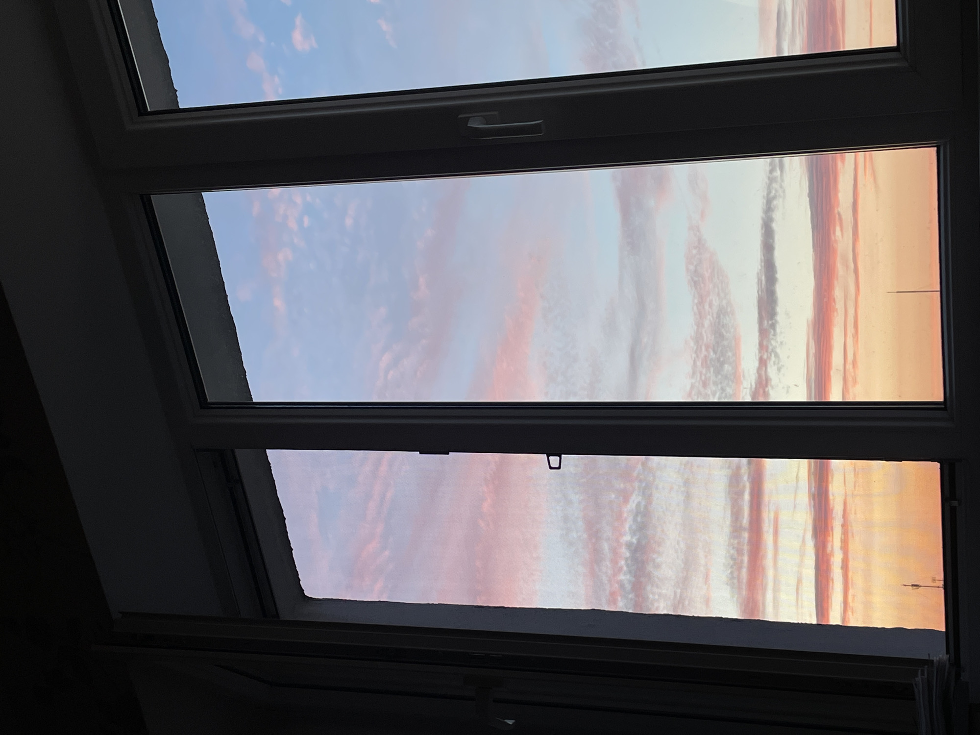 The image depicts a view of a pink and blue sky with clouds seen through a window frame. The window appears to be part of a building, with a white frame and multiple panes. One section of the window is open, revealing a darker interior space. The sky shows a gradient of colors, with warmer tones of pink and orange near the horizon, transitioning to lighter pinks and blues higher up, interspersed with wispy clouds. In the distance, silhouetted against the bright sky, are two thin vertical structures that resemble antennas or communication towers, suggesting an urban or suburban environment. The overall impression is of a sunset or sunrise viewed from an indoor vantage point. The scene is likely located in the city of Hrodna, Belarus, given the context provided.
