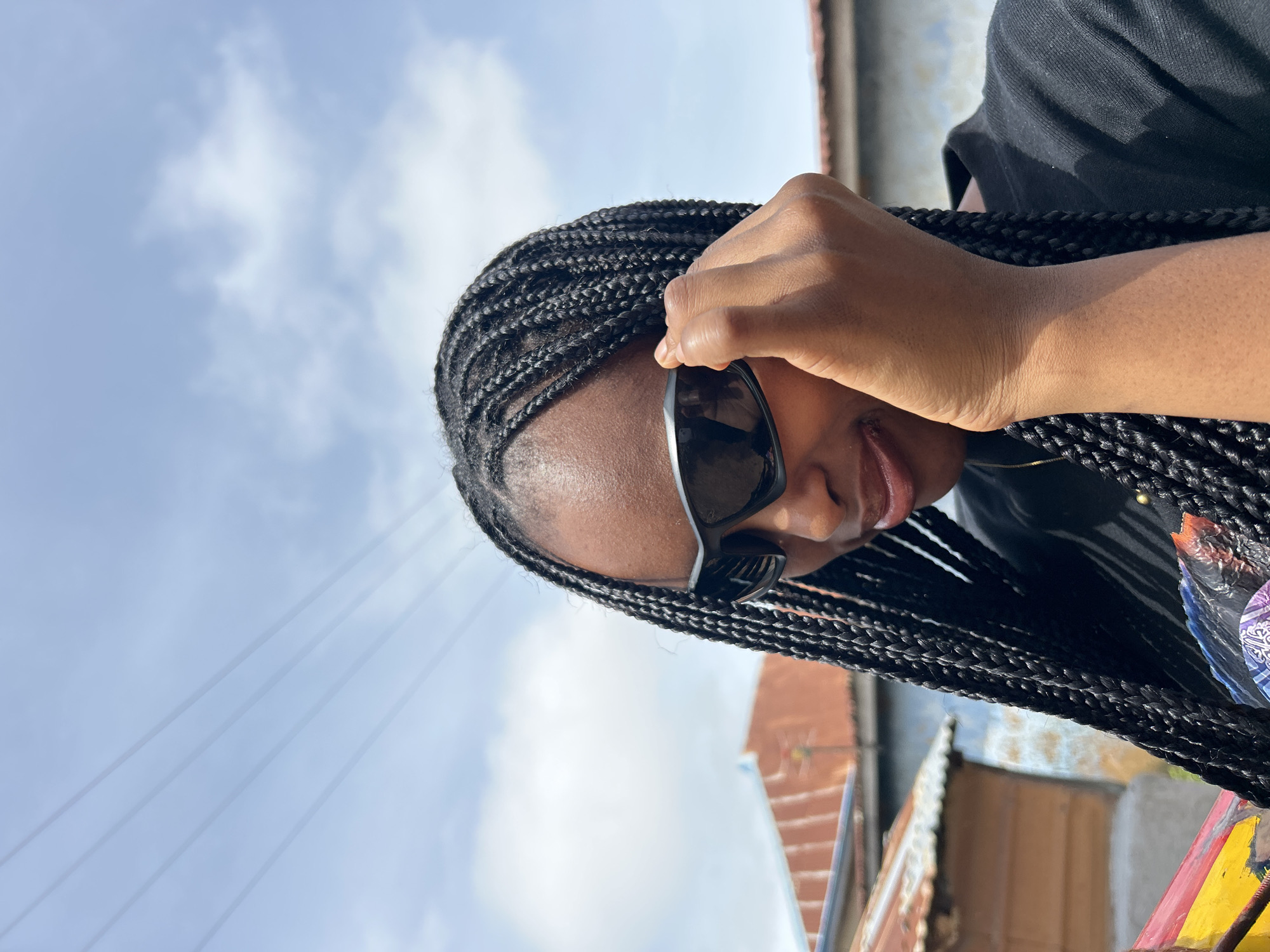 The media file is a close-up, vertically oriented photograph of a young Black person wearing sunglasses and black braids, looking downwards. The subject's right hand is raised, adjusting the sunglasses. The background consists of a bright, overcast sky with scattered clouds and some thin power lines diagonally crossing the frame from left to right. Below the sky, out of focus, are hints of buildings with reddish-brown and blue elements, possibly corrugated iron roofs and painted walls, suggesting an urban or semi-urban setting. The lighting indicates it is daytime, with the overcast sky providing soft, diffused light. The overall mood is casual and relaxed, possibly taken outdoors on a cloudy day in Jos, Nigeria. There is no visible text in the image.