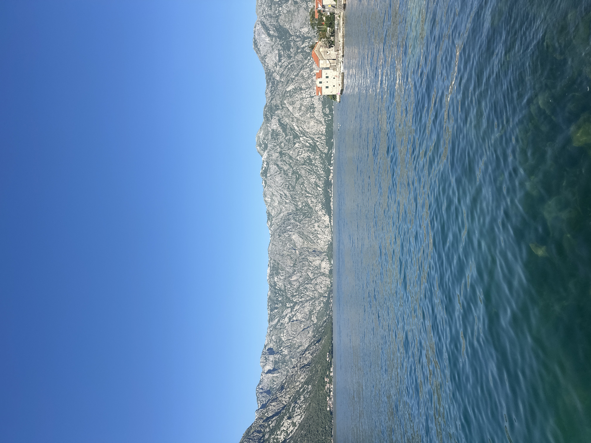 This image depicts a panoramic view of the Bay of Kotor in Stoliv, Montenegro, under a clear blue, cloudless sky that occupies the upper third of the frame. A formidable range of rugged, grey limestone mountains, partially covered with sparse green vegetation, dominates the middle ground, extending horizontally across the entire scene and rising steeply from the water. In the foreground and mid-ground, a calm body of deep blue-green water displays gentle ripples, with submerged rocks and the seabed clearly visible through the water's surface in the immediate foreground. On the right side, a small coastal settlement is visible, featuring light-colored stone buildings topped with red-tiled roofs, situated directly at the water's edge. A notable two-story building with a red roof stands on a small pier, flanked by additional structures and green foliage. Several small buoys or markers are present in the water adjacent to this settlement. Further along the distant left coastline, another, more dispersed cluster of buildings is discernible at the base of the mountains. No individuals or specific human actions are observed within the frame.
