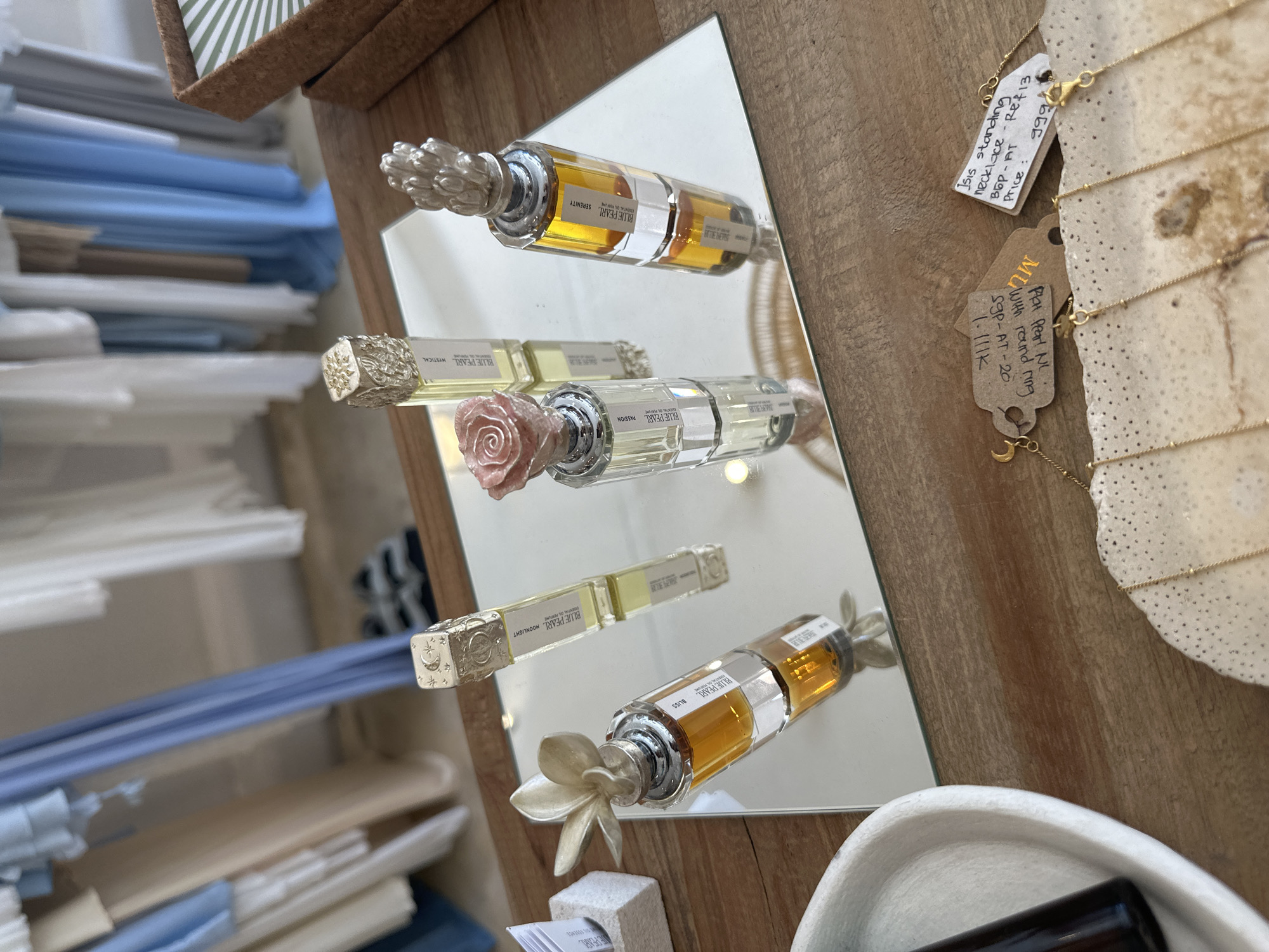 The image shows a display of "Blue Pearl" brand perfume bottles on a mirror. There are at least four bottles visible, each with different scents labeled on the bottle: "Serenity," "Passion," "Moonlight," and "Bliss." The bottles have decorative caps; one has a rose, one has a flower, and one is silver in color. The mirror reflects the bottles and the surrounding environment, including stacks of blue and white paper. A wooden surface is adjacent to the mirror, where a necklace display with price tags and annotations can be seen.
Based on the presence of the "MU" tag, it is likely this image was taken in North Kuta, Indonesia, in the area where the shop "MU" is located.