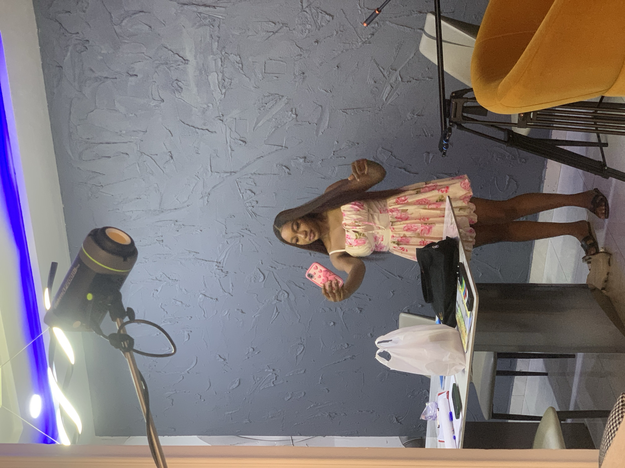 In Igboefon, Nigeria, a young adult female with long, dark hair and wearing a light-colored floral sundress stands indoors, holding a pink smartphone with a heart-patterned case at arm's length, angled as if taking a selfie or recording. Her left hand is partially raised. The background features a textured, dark gray wall. Overhead, a white ceiling with linear blue LED strip lighting and a wavy white light fixture is visible. A professional studio light, labeled "Godox SL200II," mounted on a boom arm, extends from the upper left, pointing towards the subject. To the woman's left, a white rectangular table holds a white plastic shopping bag, a black bag, a dark smartphone, papers, and a pen. A light gray chair is positioned behind the table. To her right, a black camera tripod stands. An orange upholstered armchair is visible in the lower right foreground. The floor is light-colored.