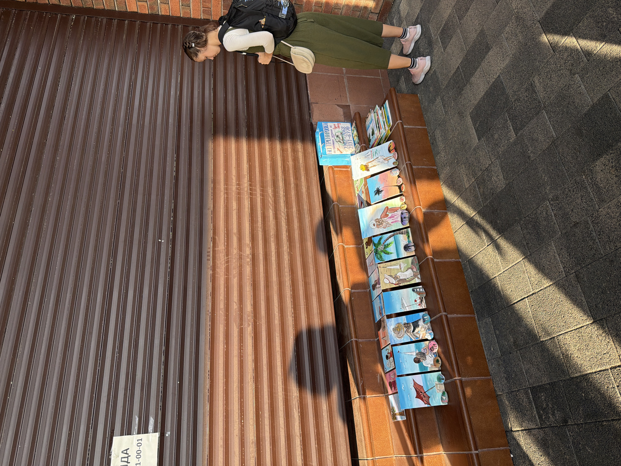 A young woman with a backpack and a crossbody bag leans over a display of colorful postcards. She is wearing a white long-sleeved shirt, olive green pants, and pink sneakers. The postcards, which appear to be illustrations of beach scenes, are arranged on a ledge. Behind the woman is a brown corrugated metal wall. The ground is paved with dark gray tiles, and sunlight casts long shadows. To the left of the woman, a small white label is affixed to the metal wall with some text. The scene suggests an outdoor market or street vendor. The lighting indicates it is daytime, likely late afternoon due to the long shadows. The backdrop of a building wall and tiled ground suggests an urban or pedestrian area.