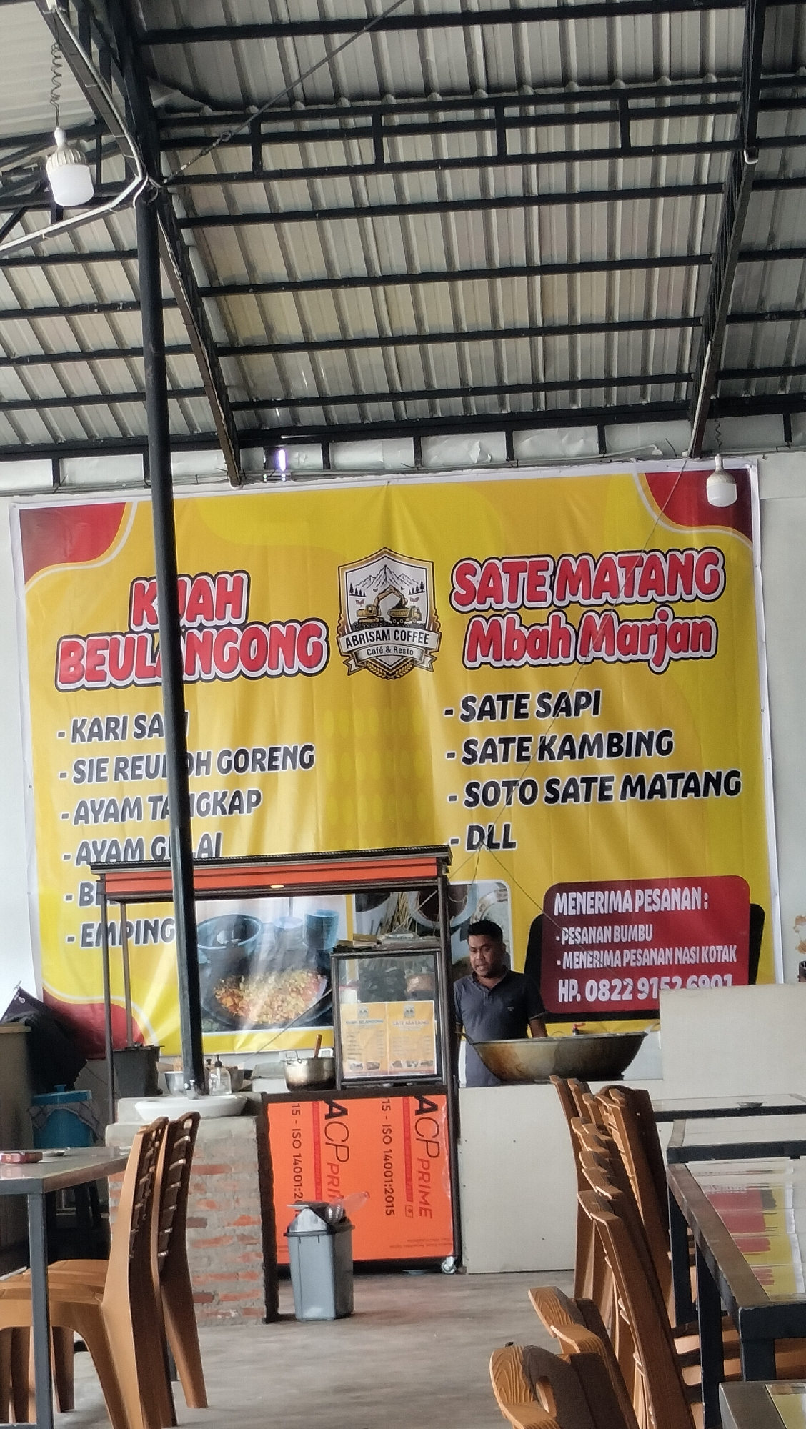 A banner with the text "SATE MATANG Mbah Marjan" hangs behind a food stall. Below the banner, various food items are listed, including "Sate Sapi" and "Soto Sate Matang". A man is standing at the stall, appearing to be cooking or serving food. In the background, there are chairs and tables, suggesting an outdoor dining area. The stall is situated in Panteraya, Indonesia.