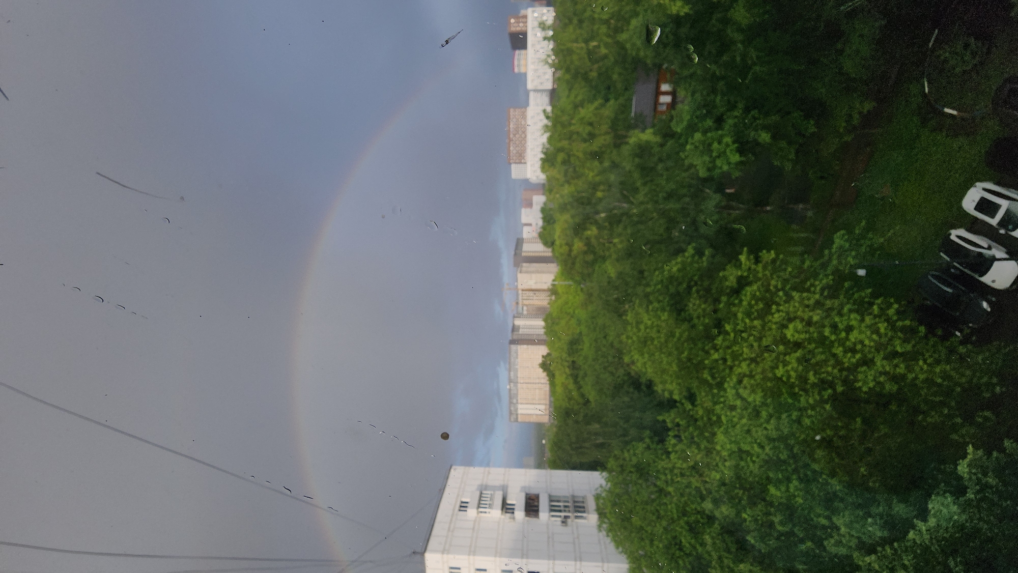 The image presents an outdoor view from an elevated perspective, likely a window, in Moscow, Russia. A partial rainbow is visible in the left portion of the frame against an overcast sky. Several apartment buildings and lush green trees dominate the middle and right sections of the image. In the lower right corner, parked cars are visible. The window through which the image was taken exhibits water droplets and streaks.