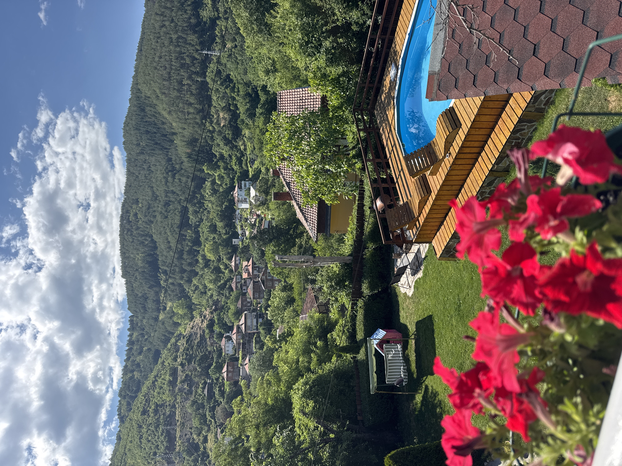 In Lilianovo, Bulgaria, a scenic residential property features a swimming pool surrounded by a wooden deck. Two lounge chairs are placed on the deck, facing the pool. In the foreground, vibrant red flowers bloom in a planter. The property also includes a green lawn with a swing set and a small playhouse. Beyond the yard, a village with red-roofed houses is nestled on a hillside covered with lush green trees. The sky above is a clear blue with scattered white clouds. A power line stretches across the upper portion of the image, and utility poles are visible on the hillside.