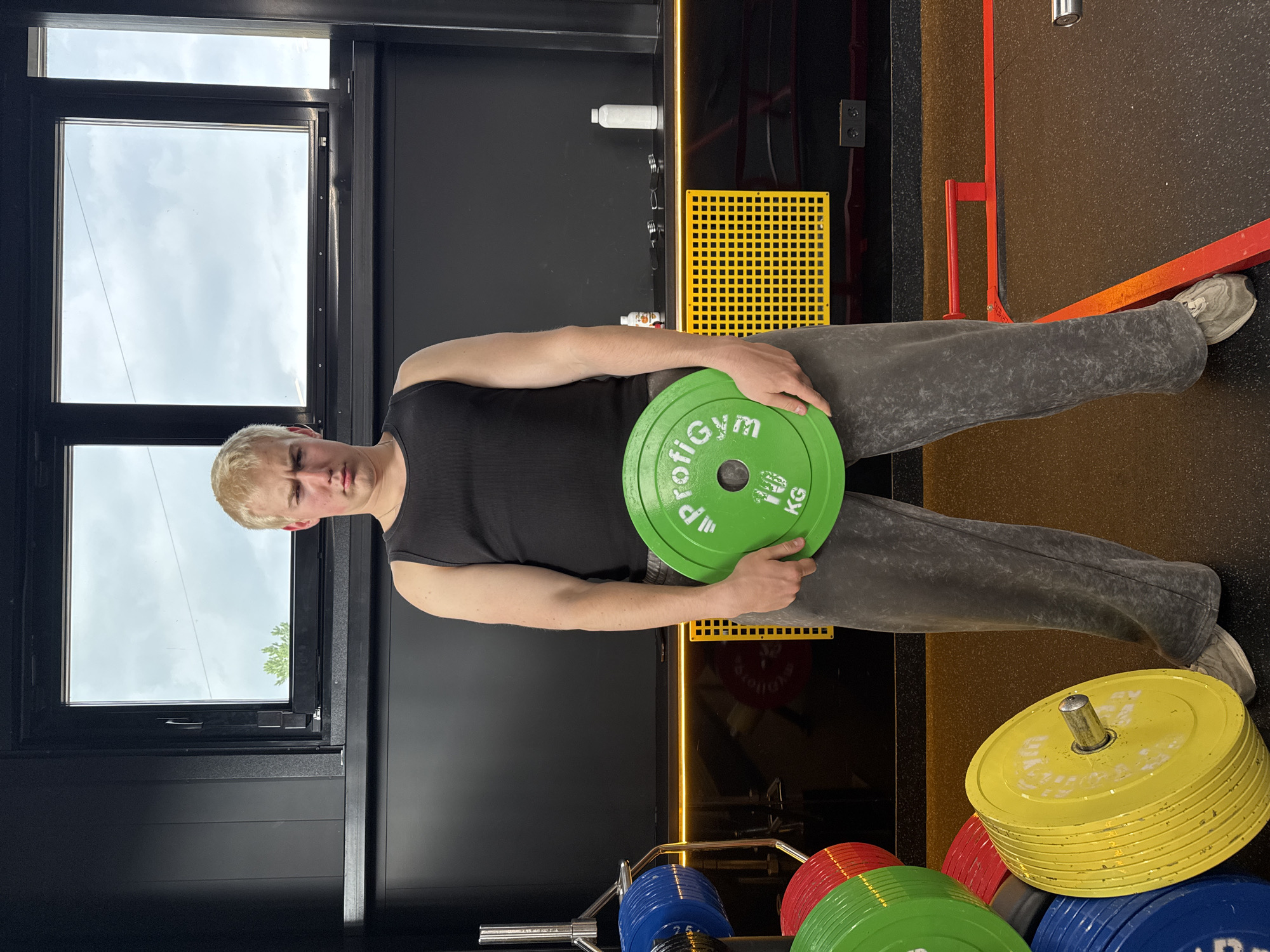 A young man with short blonde hair, wearing a black sleeveless tank top and grey sweatpants, stands holding a bright green weight plate in front of him. His expression is neutral. The weight plate he holds has "ProfiGym" and "10 KG" printed on it in white text. He is wearing light-colored sneakers.
The scene is an indoor gym or fitness center. To the man's left, a large window reveals a cloudy daytime sky, with a hint of green foliage visible. The interior features dark walls, possibly black, with a vertical orange or yellow LED light strip running along one wall. A yellow square-gridded panel is mounted on a black wall section.
In the foreground and to the right, a rack holds a variety of colored weight plates, including yellow, red, blue, and green. Red support bars are visible on the dark, textured gym floor. Other details include black electrical outlets and a white bottle or dispenser mounted on the wall. The overall impression is a modern gym environment during daylight hours.