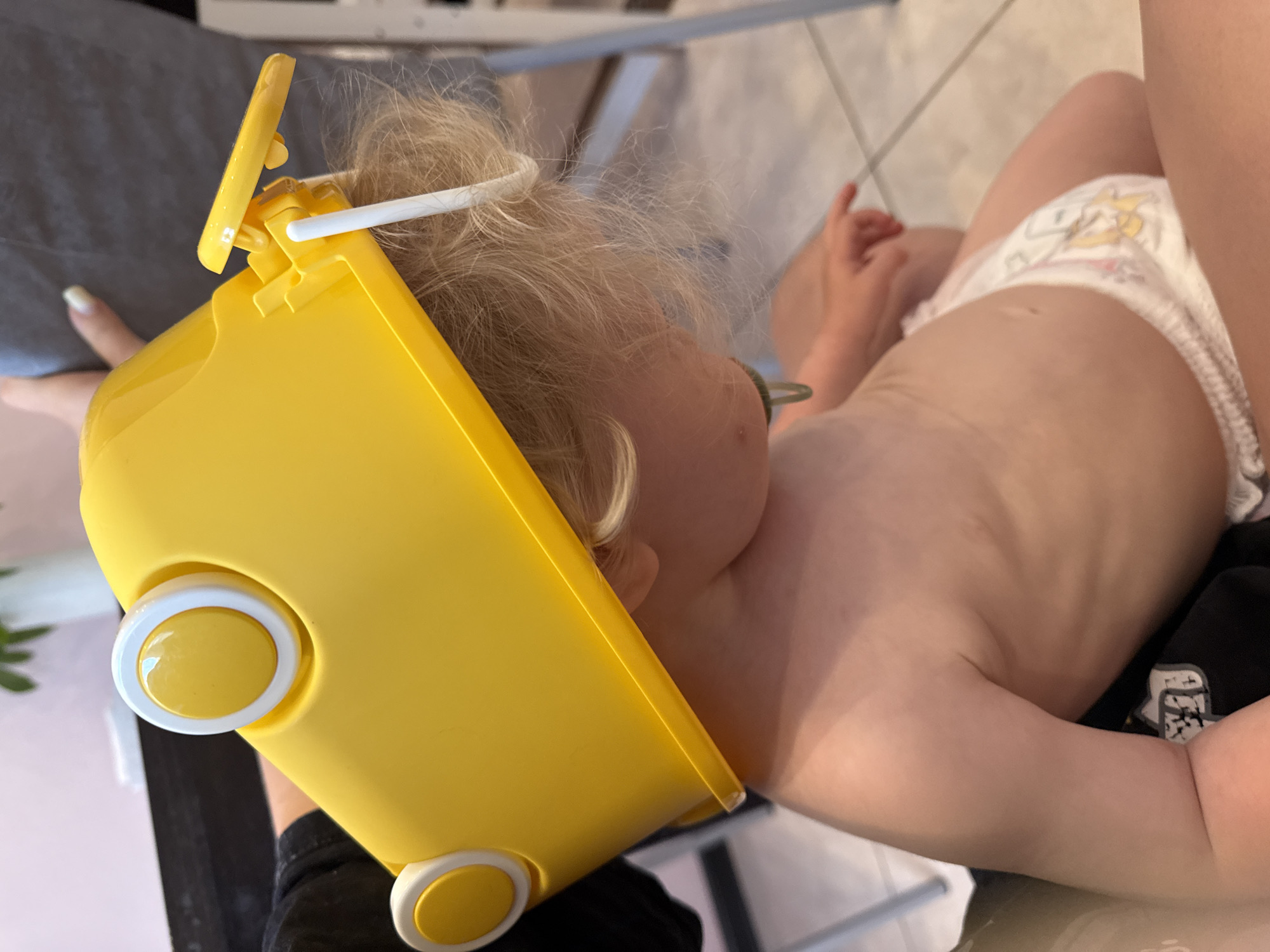 A young child with blonde hair is lying on their side, wearing a diaper and a yellow object around their head. The yellow object appears to be a helmet or a toy. The child has a pacifier in their mouth and is wearing a green cord around their neck. The scene is indoors, with a tiled floor visible in the background. The lighting suggests it is daytime. The child's expression is not clearly visible. There is no visible text in the image. The location context is Vladimir, Russia.