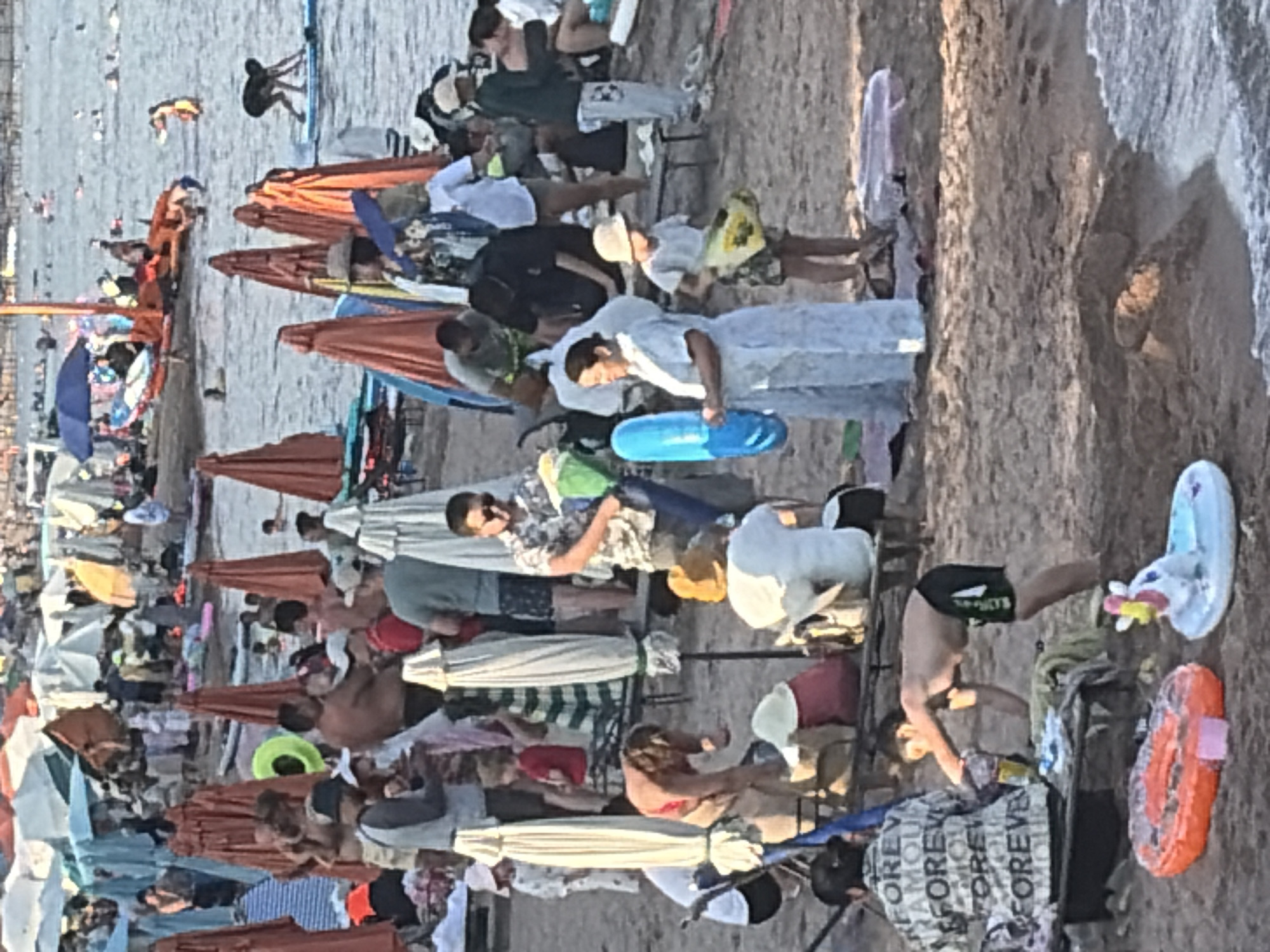 The image depicts a crowded beach scene, likely in a warm climate, with many people enjoying the water and sand. Umbrellas and beach chairs are scattered across the sand, suggesting a busy tourist destination. Several individuals are seen wading in the water, while others are relaxing on the shore. Children's toys, including inflatable rings, are visible on the beach, indicating a family-friendly atmosphere. The overall mood is one of leisure and recreation, with a sense of vibrant activity.