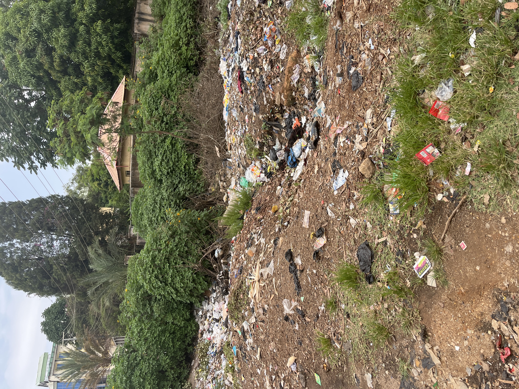 The image depicts a large, unregulated waste dump in Jos, Nigeria. The waste extends across a hillside, interspersed with patches of vegetation. Discarded materials include plastic bags, food containers, and other household refuse. In the background, buildings and trees are visible, indicating the dump is located near a residential or commercial area. There is no presence of people or animals within the image.