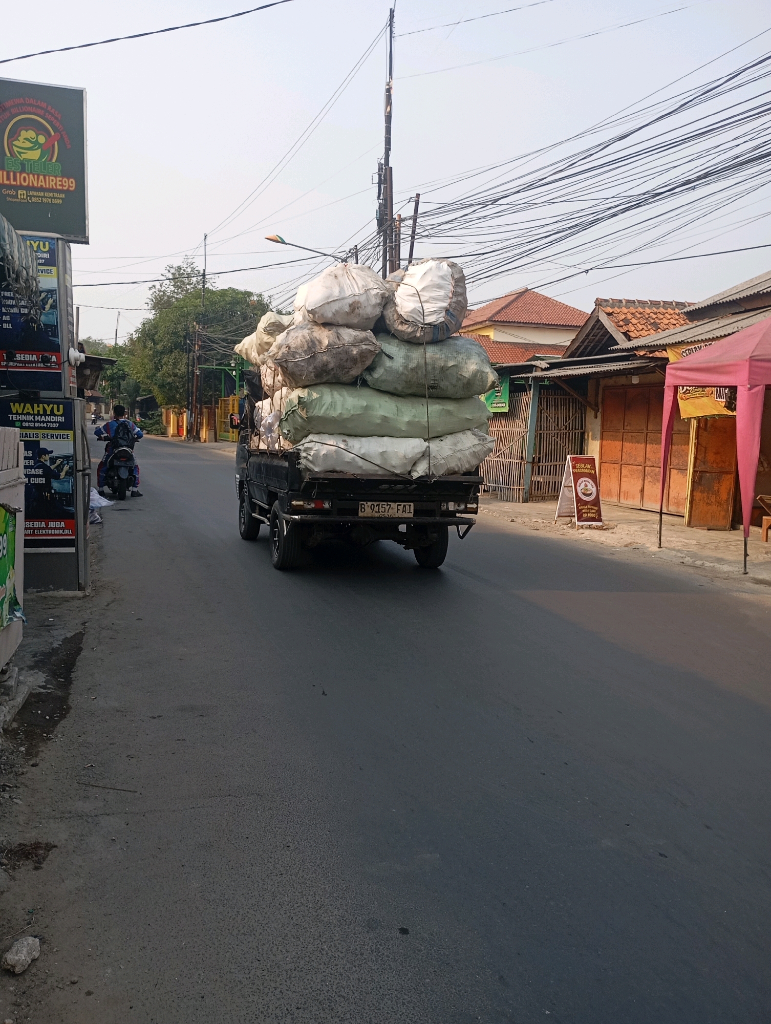 In Babelan, Indonesia, a black pickup truck with a license plate reading "B 9157 FAI" is driving down a road. The truck is heavily loaded with large sacks of what appears to be goods, stacked high. On the left side of the road, a person on a motorcycle is facing away from the camera. Several buildings line the right side of the road, including one with a pink awning. Overhead, a dense network of power lines is visible against a hazy sky. A sign on the left also advertises "ES TELLER MILLIONAIRE99."