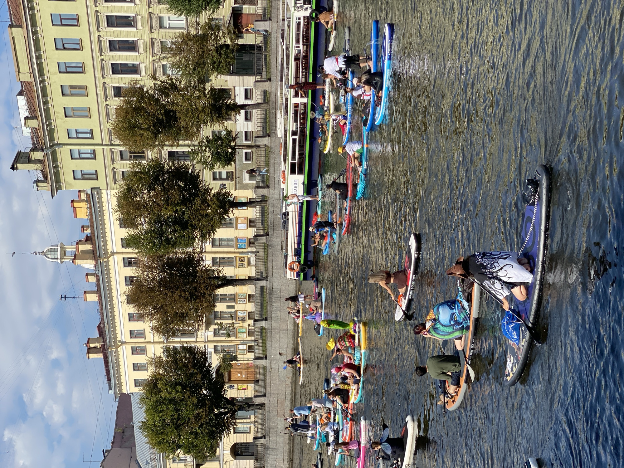 This image depicts a large group of people paddleboarding on a canal in Saint Petersburg, Russia. The canal is flanked by historic buildings with classical architecture and lined with trees. The water is calm, and the sky is partly cloudy. Numerous individuals are spread across the canal, some standing on their paddleboards and others sitting or kneeling. Many are holding paddles, and some are engaged in conversation. A large passenger boat is visible in the background, as are several bridges and other structures typical of the city. The scene appears to be a recreational event or gathering, possibly a themed paddleboarding excursion given the variety of attire and accessories among the participants. The overall atmosphere is lively and communal, with a focus on outdoor activity in an urban waterfront setting.