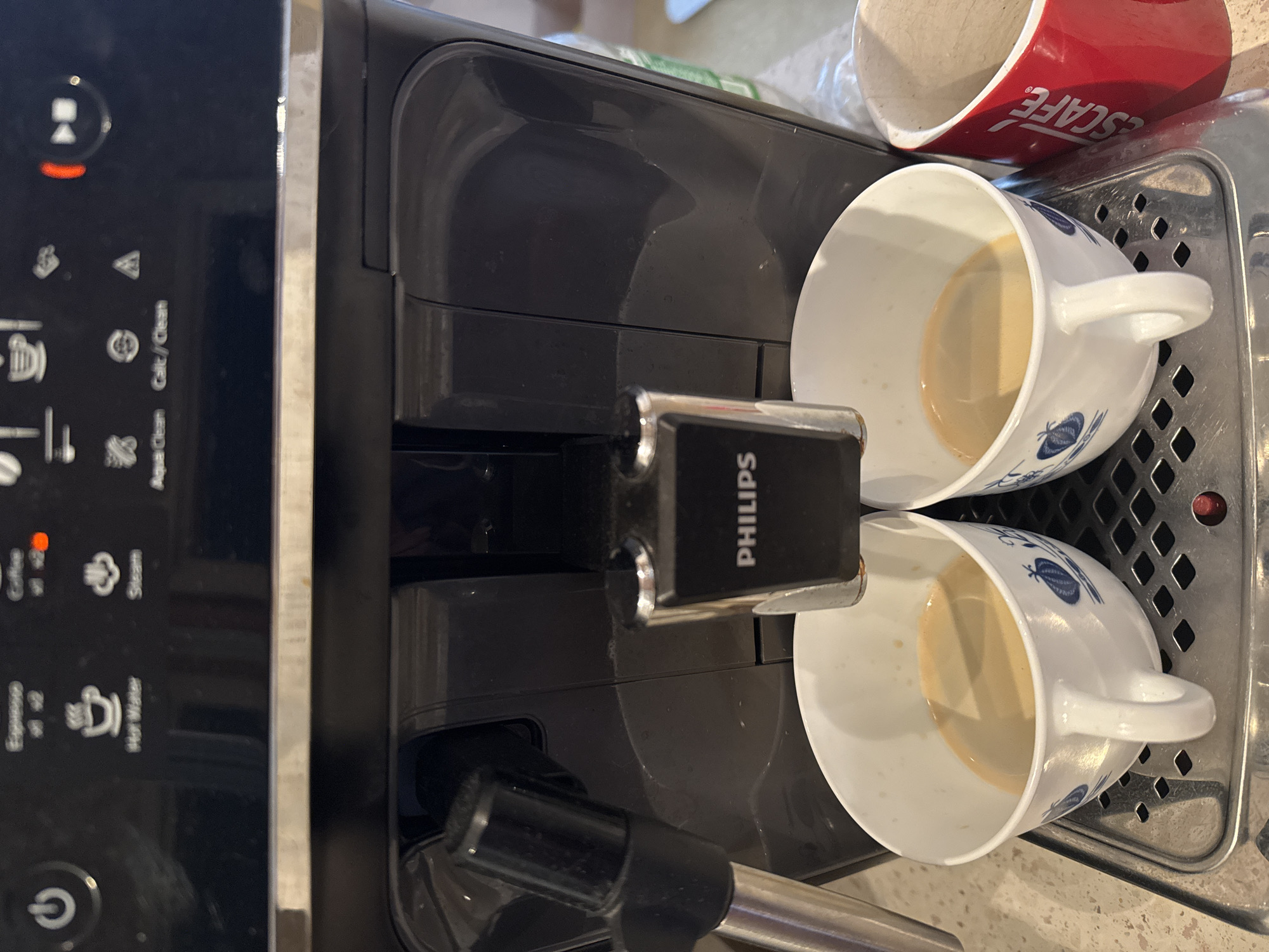 Here is a detailed description of the image:
**Scene Description:**
The image shows a Philips espresso machine dispensing coffee into two white cups with blue floral designs. The machine has a black control panel with various illuminated icons. The machine is dispensing coffee into two cups placed on the drip tray. A red "ESCAPE" branded coffee cup is also seen in the shot. The scene seems to be set in a kitchen or office break room. The exact location in the city of Unknown, Unknown is not possible to determine from this image alone.
**Objects:**
* Philips espresso machine
* Two white cups with blue floral designs
* A red "ESCAPE" coffee cup
* Stainless steel drip tray
**Actions:**
* Espresso machine dispensing coffee
**Environment:**
* Interior setting, likely a kitchen or office break room
**Details:**
* The cups appear to be partially filled with coffee.
* The Philips brand name is visible on the dispensing arm.