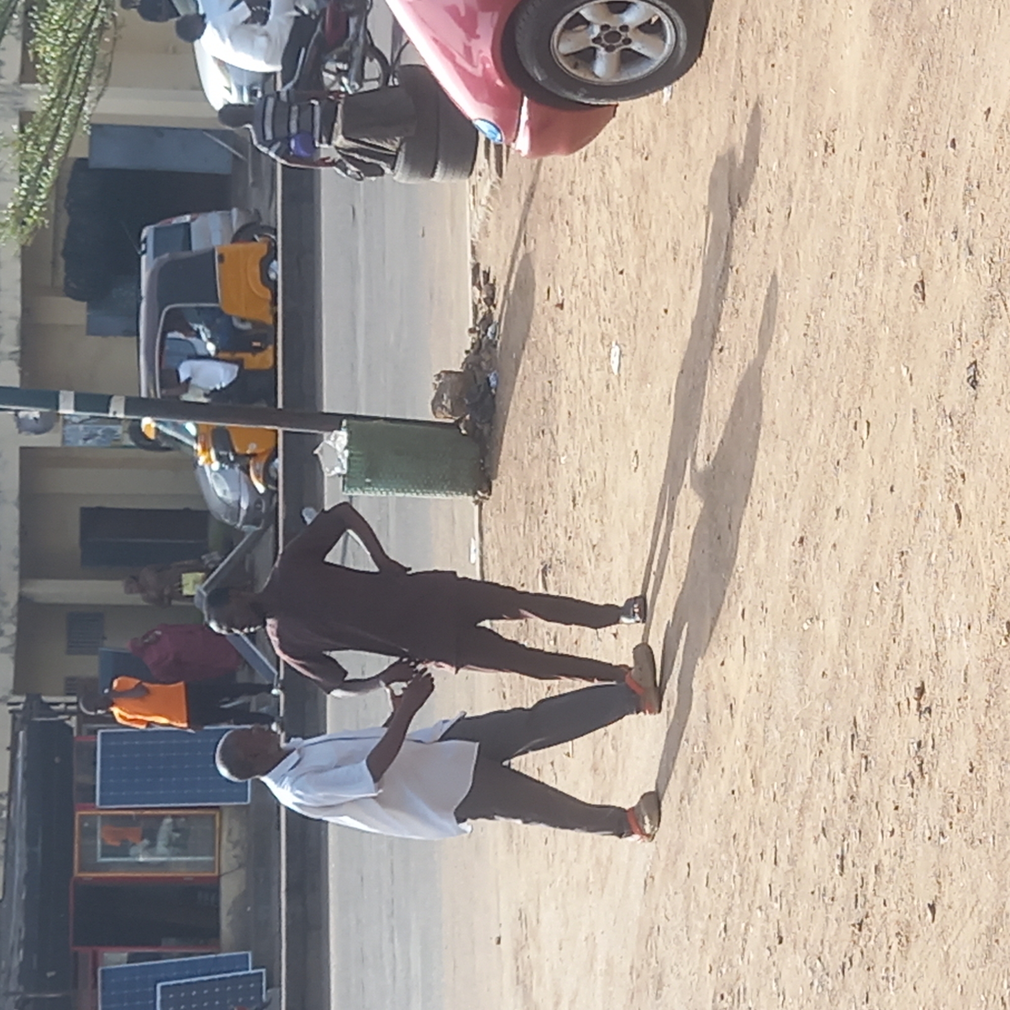 This media file captures a sunlit street scene in Dadi Riba, Nigeria, likely during late afternoon or early morning due to the long shadows cast. The ground on the right side of the image is unpaved and sandy, while a paved road or sidewalk runs along the left, bordered by buildings.
In the foreground, two men are walking or standing on the dirt path. One man wears a dark shirt and dark trousers, standing with his hands on his hips. The other, in a white shirt and dark trousers, is positioned slightly in front, also moving towards the right. Both cast prominent, elongated shadows across the sandy ground.
Between the dirt path and the paved road, a green metal trash bin is visible, surrounded by some debris. A red car is parked behind the bin, with part of its rear visible. Nearby, several motorcycles or scooters, including a prominent white one, are also parked.
Further back on the paved road, a yellow Keke Napep (tricycle taxi) is present, with people seated inside. Other individuals are visible in the background, including a person in a red shirt and another in an orange shirt, near what appear to be commercial establishments. On the far left, several blue solar panels are displayed upright, suggesting a vendor or an area for selling electrical goods, alongside what might be televisions or other appliances.
The scene conveys a typical busy street environment with local transportation, casual interactions, and commercial activity. No legible text is visible in the image.