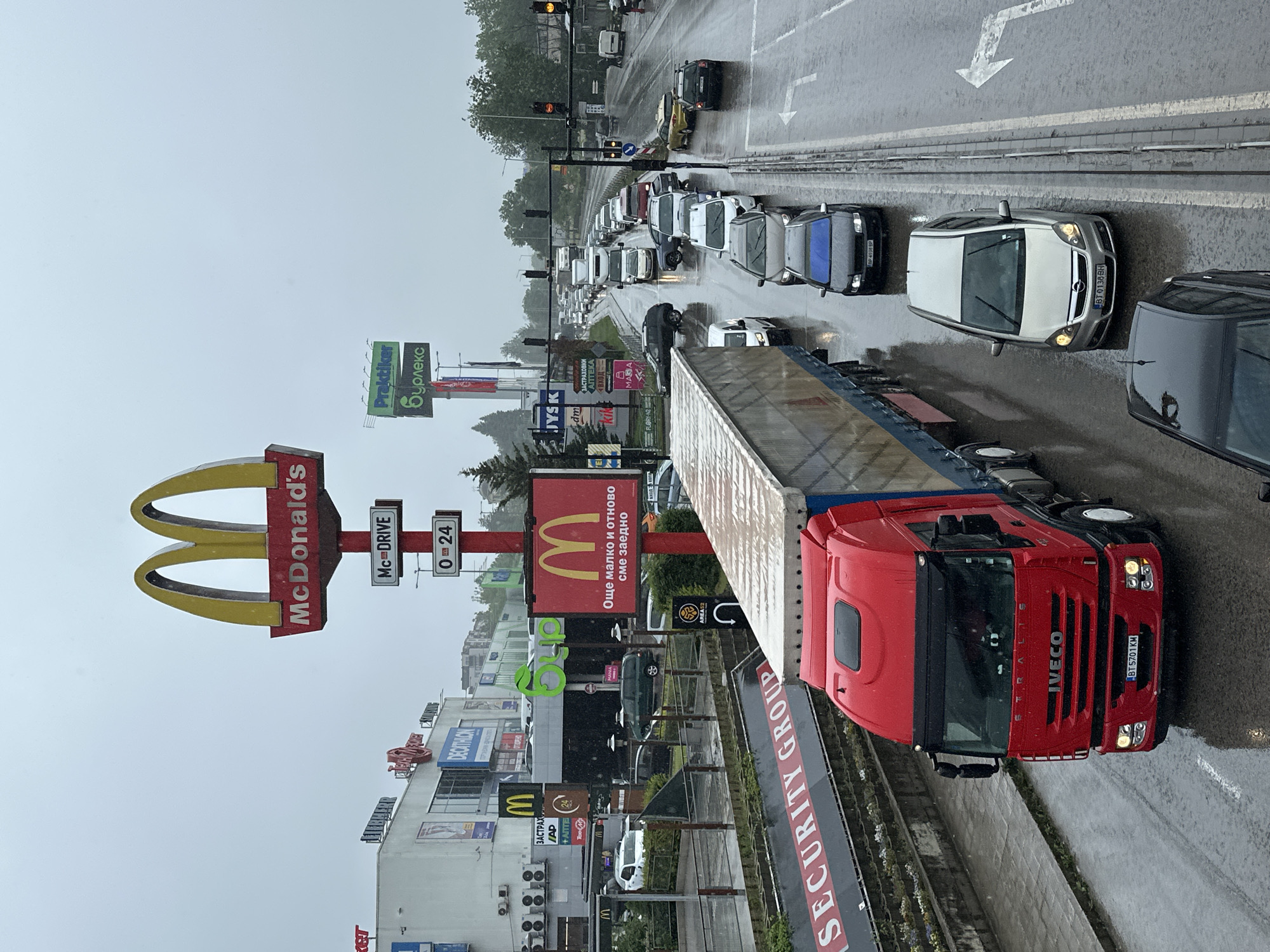 The image shows a traffic jam in Veliko Tarnovo, Bulgaria, on a rainy day. A red IVECO truck dominates the foreground, positioned next to a "SECURITY GROUP" sign and a guardrail. In the background, numerous cars are congested on the road. Prominent signs for businesses, including McDonald's (with "Mc-DRIVE" and "0-24" hour signage), Praktiker, and Decathlon, are visible on the left, indicating a commercial district. A smaller sign for JYSK is also present. The McDonald's sign in Cyrillic states "Още малко и отново сме заедно" (a little more and we are together again). Other signs and businesses are visible, including dm, Apteka, and Area 52. There are traffic lights ahead and street markings indicate lane direction. The weather appears overcast and wet. The license plate number of the Opel Astra in the rightmost lane is BT0138BH, and the truck's license plate is BT5701KM.