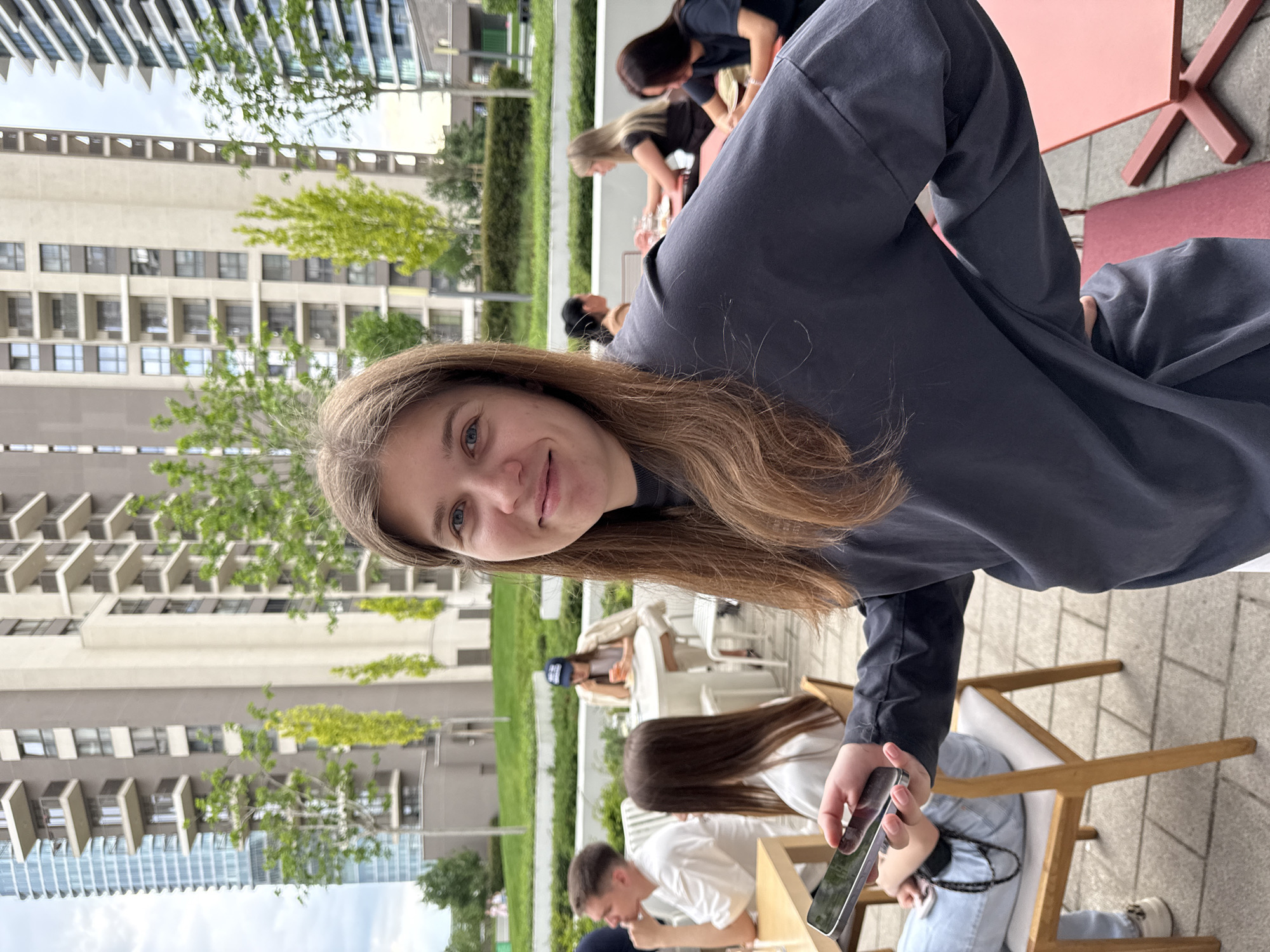 The image depicts a young woman with long brown hair, wearing a dark blue long-sleeved shirt, holding a smartphone and smiling at the camera. She is seated outdoors, likely at a cafe or restaurant. Behind her are other people, including a group sitting at a table, and some walking.
The background shows a modern urban environment, with tall buildings and greenery, suggesting a city location. Based on other images, the location is in Belgrade, Serbia. The buildings are of modern design, with multiple windows and balconies. There are also trees and shrubs visible, indicating a landscaped area. The setting appears to be a public space or outdoor dining area.