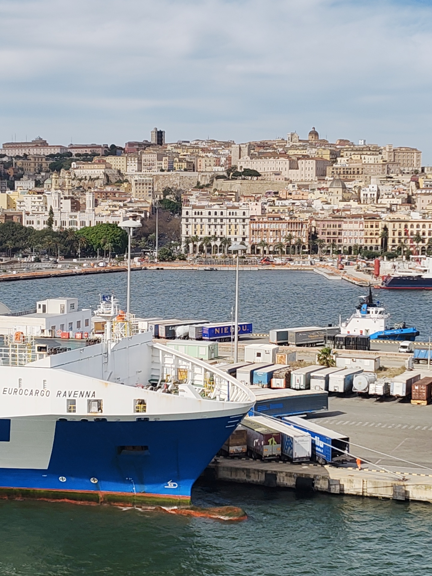 The image captures a harbor scene in Cagliari, Italy. In the foreground, the bow of the EUROCARGO RAVENNA, a blue and white cargo ship, dominates the view, docked at a pier. A variety of trailers and shipping containers are lined up on the pier, suggesting active loading or unloading operations. An individual in an orange vest is seen near the pier edge. In the background, the city of Cagliari rises in tiers, with historical buildings and landmarks visible against a slightly overcast sky. The waters of the harbor are visible between the ship and the shoreline, with a tugboat positioned nearby.