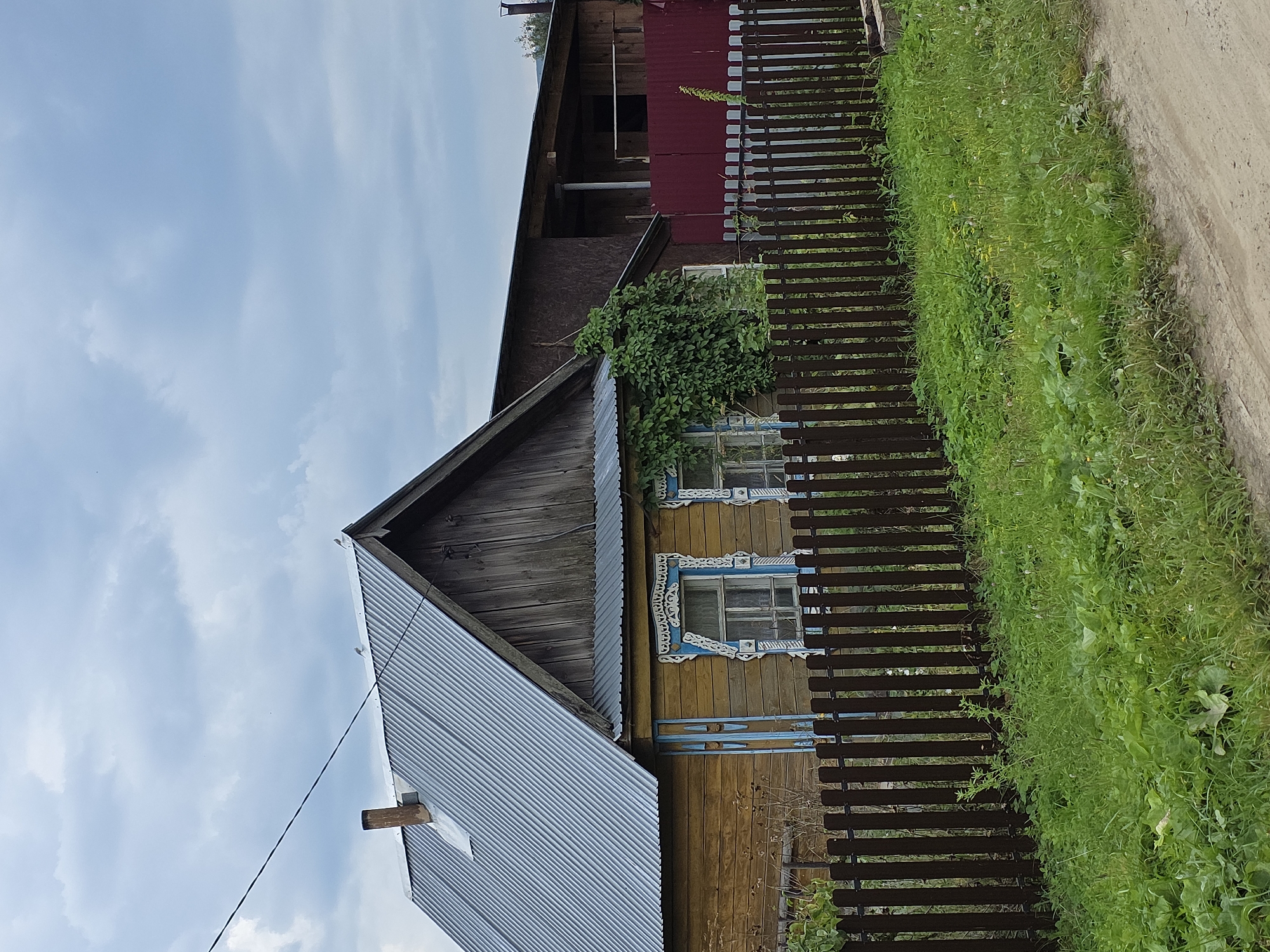 The image shows a wooden house with a corrugated metal roof and decorative window frames in Magnitka, Russia. A dark wooden fence runs along the front of the property, with overgrown green grass and vegetation behind it. The sky is cloudy. To the right of the house is a darker, larger building, partially obscured by greenery, and a red fence. A dirt road is visible in the foreground.