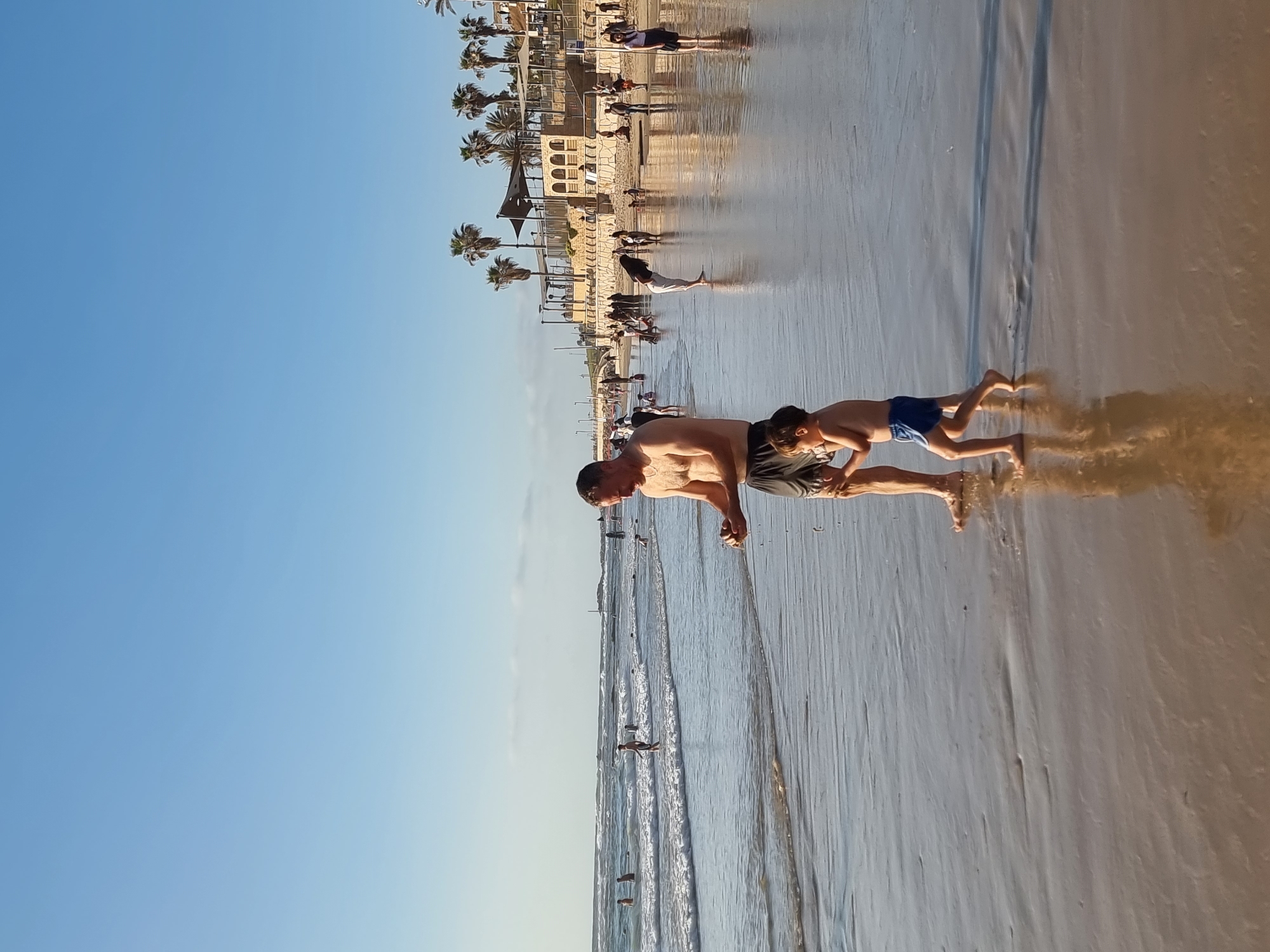 The image depicts a man and a child wading in the shallow surf of a beach. The man, shirtless and wearing dark shorts, is partially bent over, appearing to interact with the child. The child, wearing blue swimming trunks, is farther into the water. The water is shallow and reflects the sky, with sand visible underneath. The beach is crowded with many people. In the background, a line of palm trees and buildings is visible, suggesting the beachfront of Tel Aviv, Israel. The sky is clear and blue.