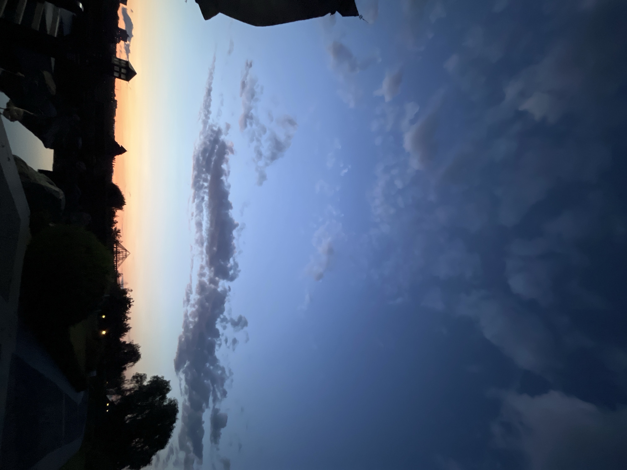 The image depicts a silhouetted landscape at dusk. The sky displays a gradient of deep blue at the top transitioning to lighter blues and oranges near the horizon. Scattered clouds are present in the sky. In the foreground, dark shapes suggest a natural or landscaped area with what appears to be water or a reflective surface. Silhouetted structures, including what might be architectural elements or art installations resembling tilted buildings, are visible on the left. Further back, a treeline separates the foreground from the horizon, with some lights faintly illuminating the scene. The overall impression is a park or outdoor recreational area. The mention of Dnipro, Ukraine, suggests this location is within that city, though specific identifiable landmarks that confirm this are not apparent from the image itself. The scene is devoid of any visible people.