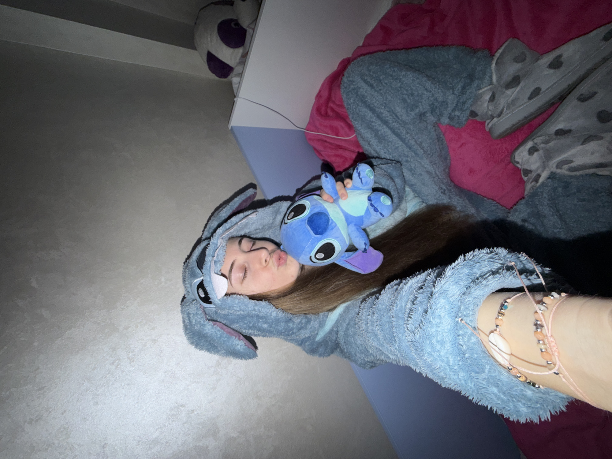 A girl in a blue "Stitch" onesie is lying on a bed, holding a "Stitch" plush toy. She has bracelets on her wrist. There is a pink blanket on the bed and a panda plushie in the background. The location cannot be determined from the image.