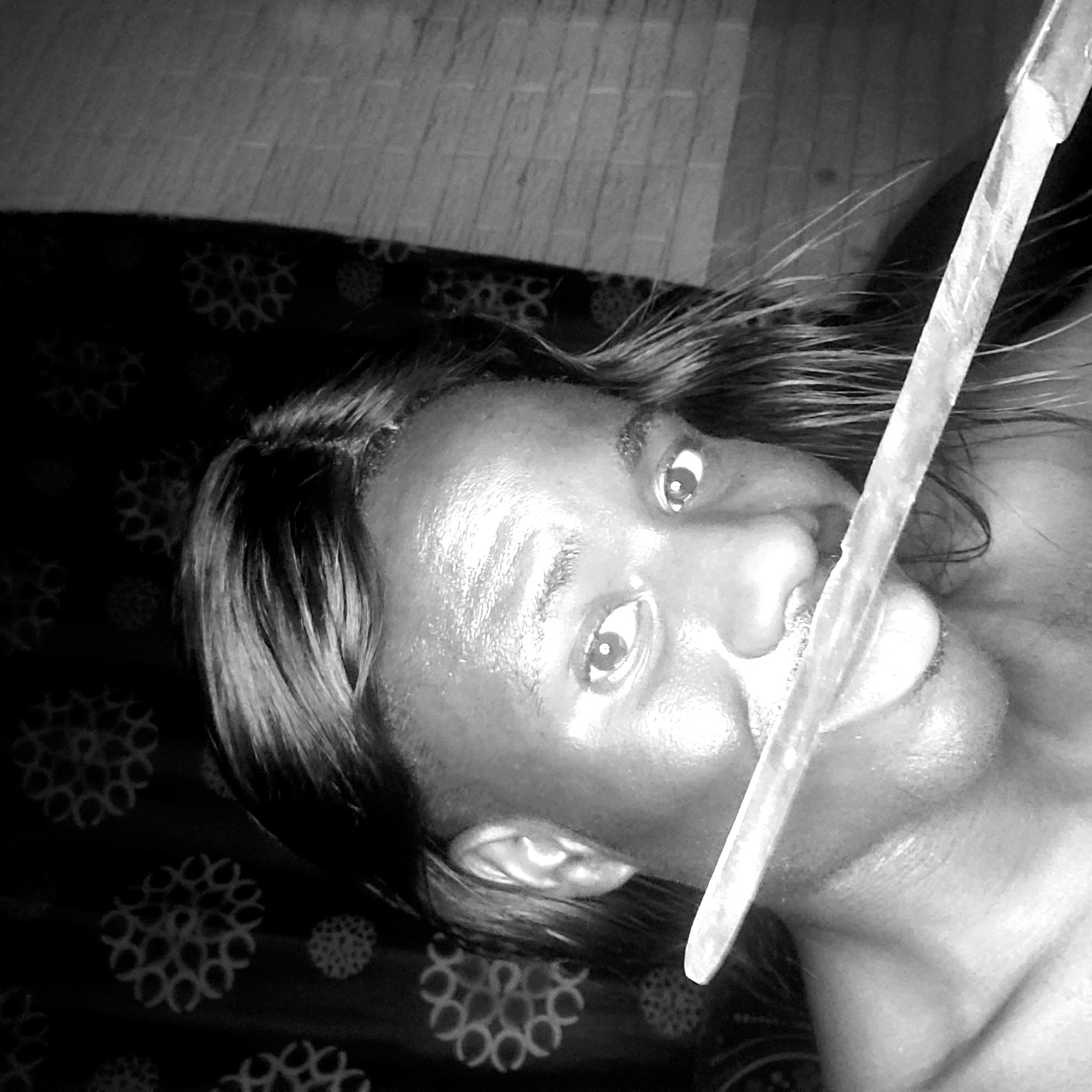 A close-up, black and white photograph shows a person with dark skin and long, dark hair lying on a patterned fabric. The person's face is angled upwards, with wide eyes looking directly at the camera. A straight, light-colored object, possibly a blade, is positioned vertically across the person's face, from forehead to chin. The background includes patterned fabric and a section of what appears to be wooden flooring. The location is not evident based on the image.