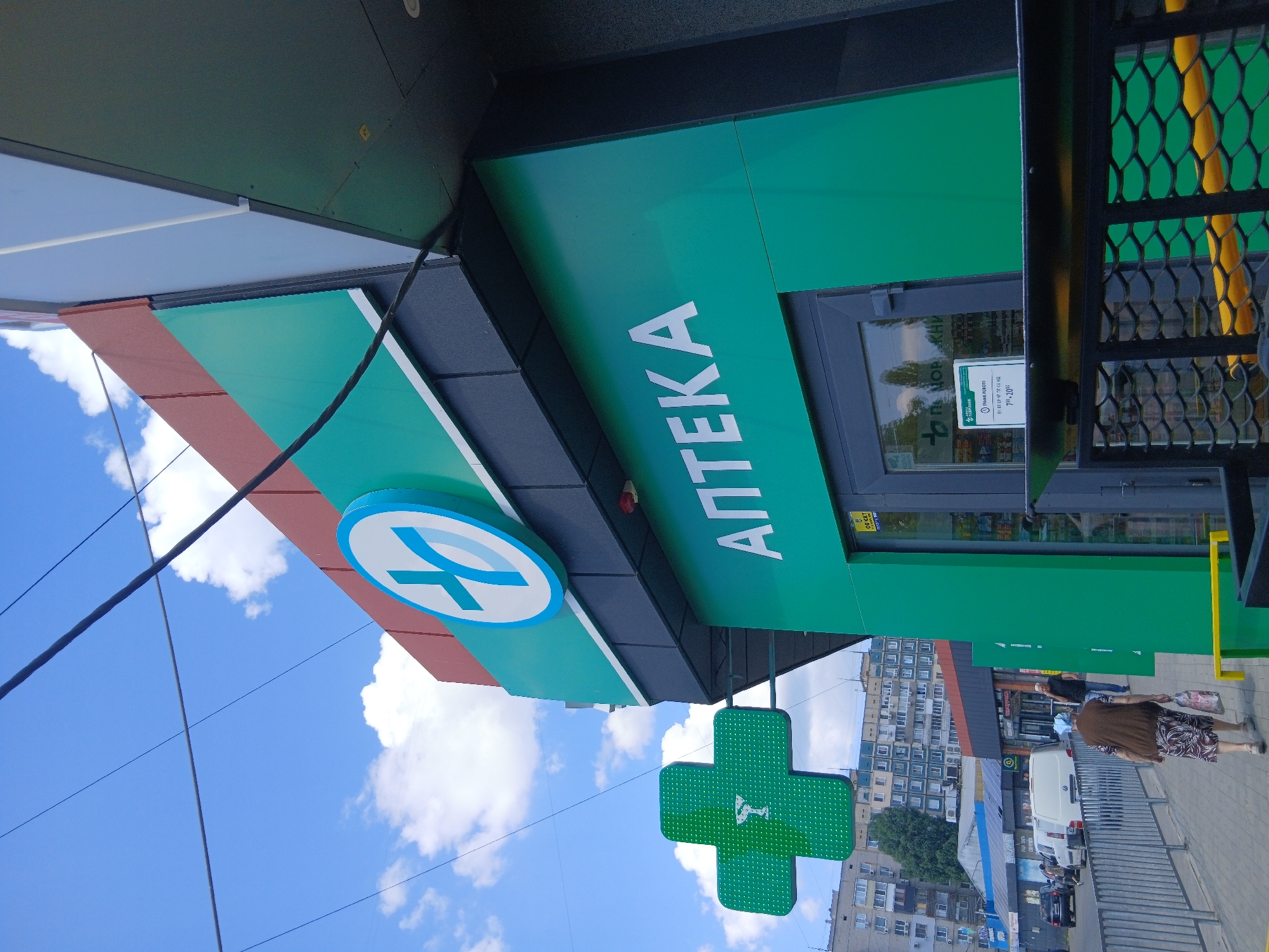 The image shows a pharmacy with a bright green facade and large white lettering that reads "АПТЕКА" (Apteka, meaning Pharmacy). Above the entrance, a circular sign with a blue cross inside a white circle is visible. To the left of the building, a prominent green cross sign with a medical symbol is illuminated. The scene is outdoors, under a blue sky with scattered white clouds. Several people are walking on the sidewalk in front of the pharmacy. One person, an elderly woman wearing a headscarf and a brown vest over a patterned dress, is walking away from the camera carrying a shopping bag. Another person, a man in a dark shirt, is walking in the opposite direction. In the background, there are apartment buildings and other shops. A fence runs along the sidewalk. The image appears to have been taken on a sunny day. Based on the signage and architectural style, the location is likely in the city of Dnipro, Ukraine.