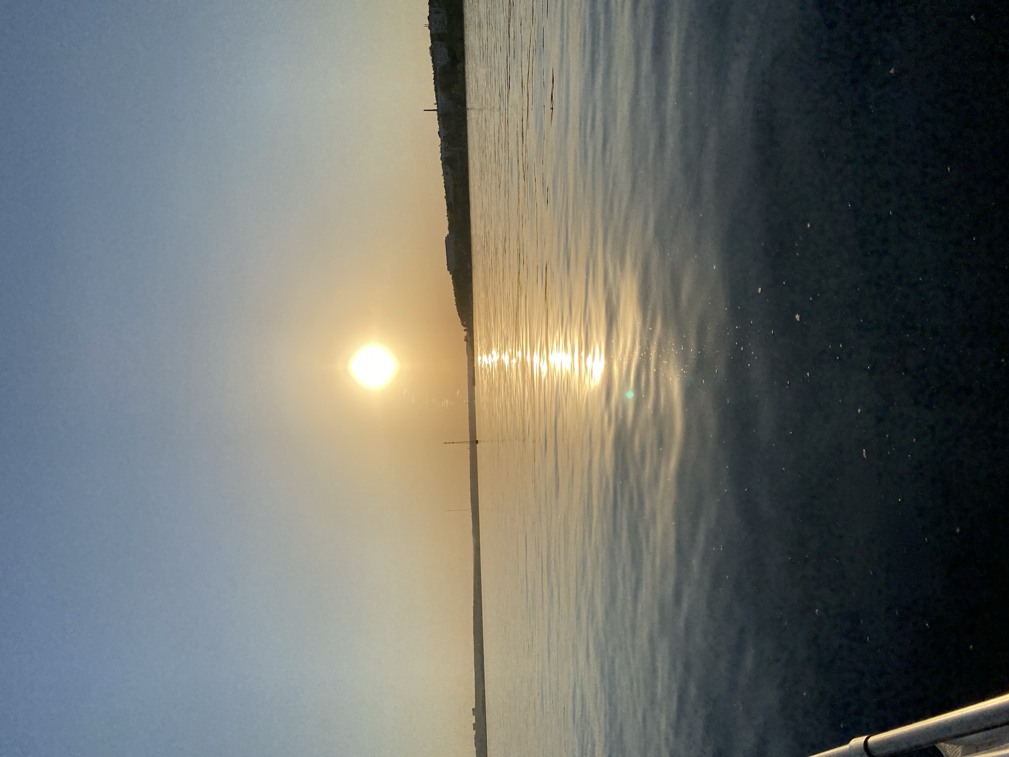 The image depicts a body of water, likely a river or large lake, with the sun reflecting off the surface. The sun is positioned on the left side of the frame, and a line of bright reflection extends across the water towards the right. On the opposite shore, there are buildings and potentially some industrial structures. The image is somewhat hazy, likely due to atmospheric conditions. The bottom corner shows a railing. The location is likely the Izhevsk pond.