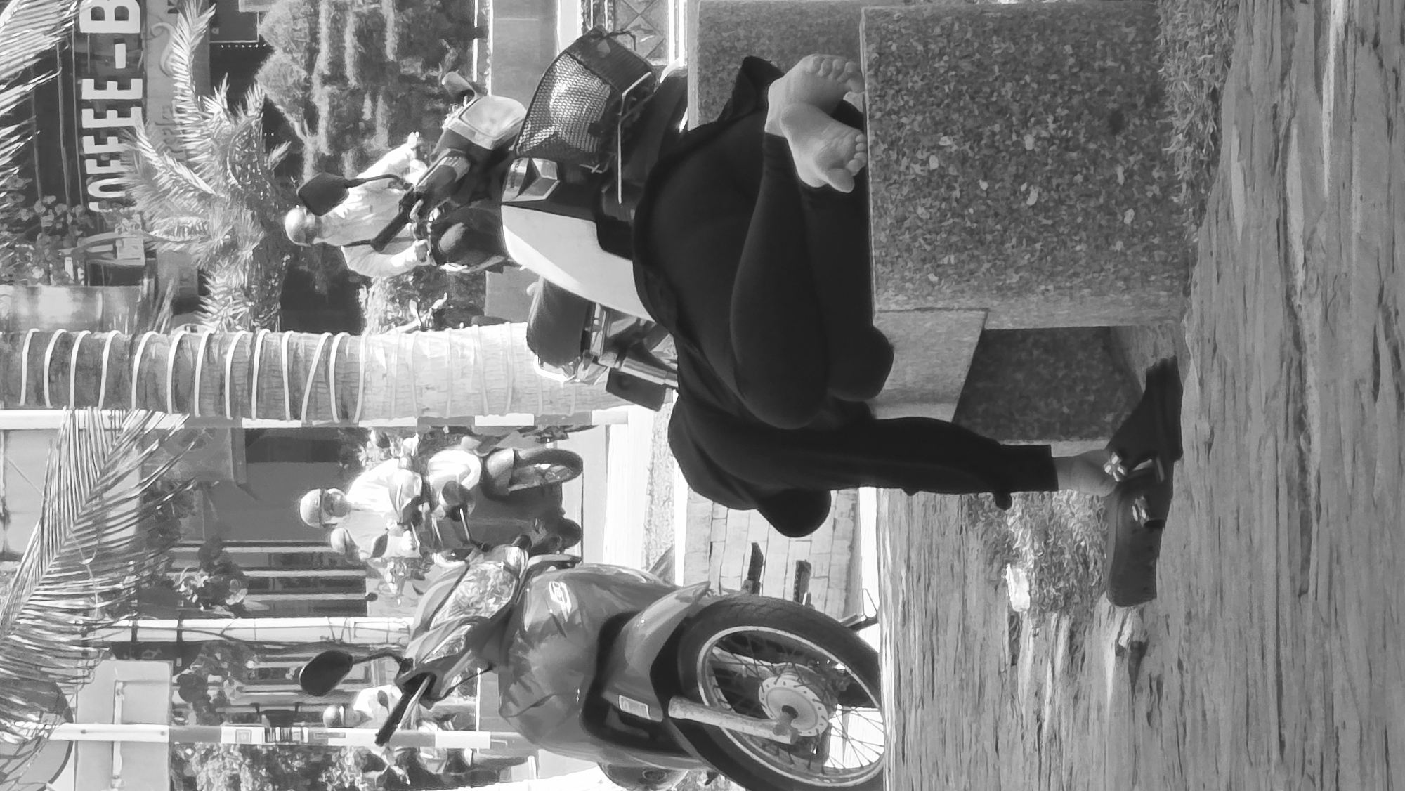The image shows a street scene in Nha Trang, Vietnam. A person is sitting on a bench with their bare feet resting on the back of the bench. A motorcycle is parked in front of the bench. Another person on a motorcycle is driving by in the background. A building with a sign that says "COFFEE - B" is visible in the background. A palm tree is also visible.