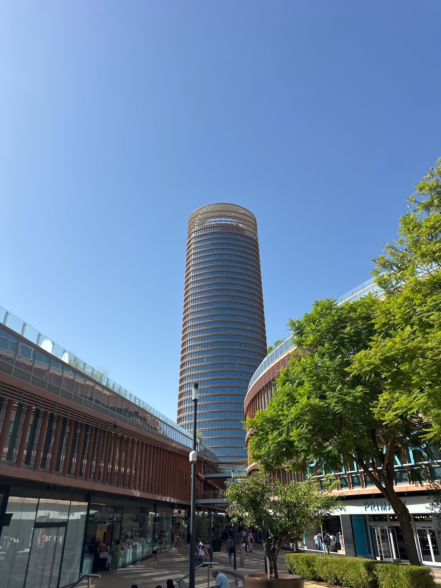 The image captures a street-level view of the architectural complex in Seville, Spain, featuring the iconic Torre Sevilla skyscraper and a modern shopping center with curved facades. The Torre Sevilla, a tall, cylindrical building with a distinctive brown lattice exterior and numerous glass windows, dominates the background under a clear blue sky. In the foreground, the shopping center exhibits a contemporary design with copper-colored louvers and glass railings, reflecting the sky and surrounding greenery. Lush green trees are strategically placed, softening the urban landscape and providing shade. Several shops are visible, including "Primark," with its entrance featuring automatic glass doors. People are walking along the pedestrian area, some browsing shop windows, and others seated on benches, indicating a public space with commercial activity. The overall scene depicts a vibrant and modern urban environment within Seville.