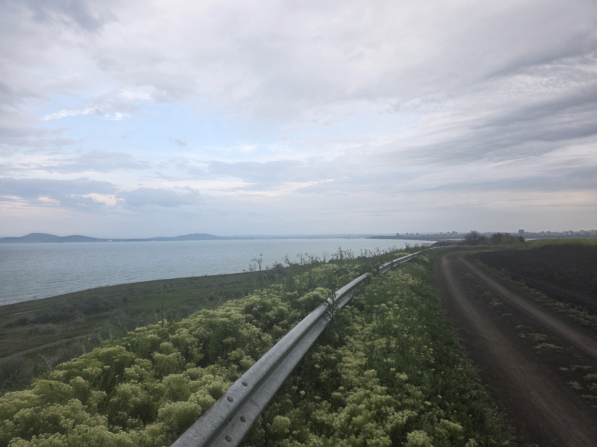 The image shows a coastal scene in Burgas, Bulgaria. A dirt road runs along the edge of a field, bordered by a metal guardrail and dense vegetation. Beyond the vegetation is the Black Sea, with land visible in the distance. The sky is overcast with grey and white clouds. No people are visible in the scene.
