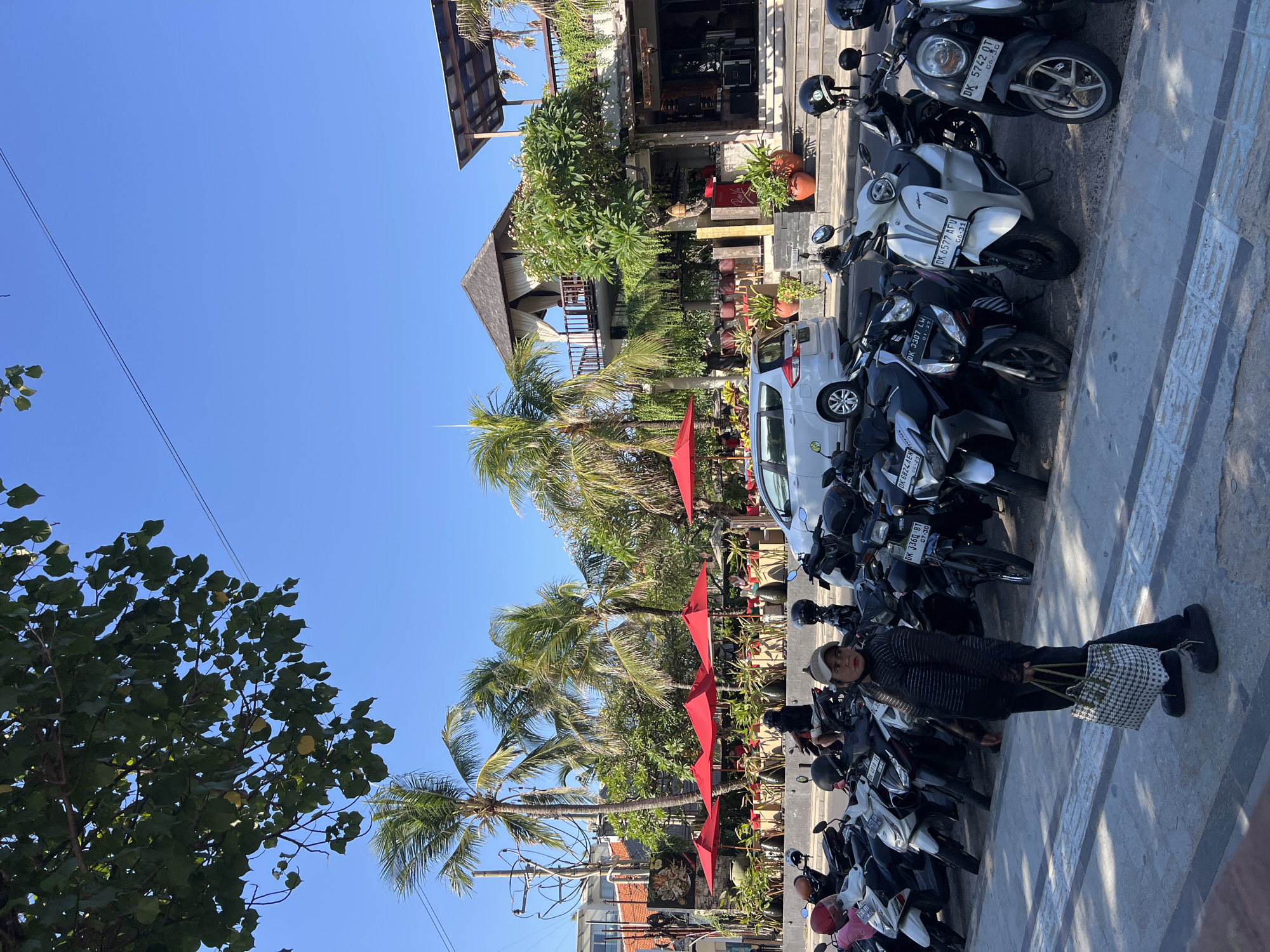 The image captures a street scene in Kuta, Indonesia, on a sunny day with a clear blue sky. The foreground features a row of parked motorcycles, primarily scooters, in various colors including black, white, and silver. A person wearing dark clothing and a cap stands on the sidewalk, holding a woven bag. Behind the motorcycles, a multi-story building with an open-air second-floor balcony is visible. The building is adorned with lush greenery and has an entrance area with red stools and potted plants. Red umbrellas are set up in an outdoor seating area in front of the building, suggesting a restaurant or cafe. Palm trees are scattered throughout the scene, framing the architecture and adding to the tropical ambiance. A white car is parked on the street adjacent to the motorcycles. The ground is paved with a textured walkway in the foreground and a darker asphalt road. Overhead, power lines are visible against the sky.
