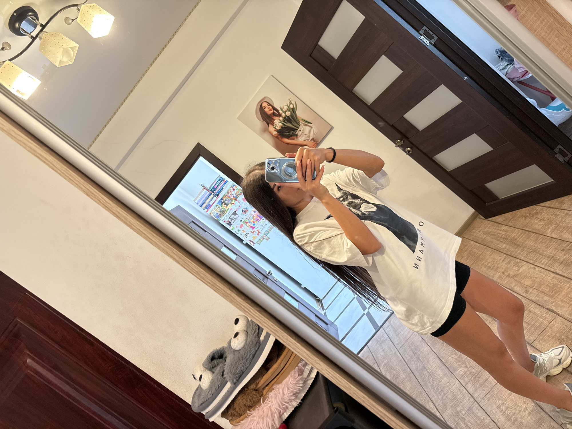 In a well-lit room, a young woman with long dark hair is taking a mirror selfie. She is wearing a loose white t-shirt with a graphic print and black shorts, paired with athletic shoes. The room features a large mirror on the left, reflecting a dark wooden door and a wall with a framed picture of a woman holding tulips. A stack of fluffy slippers is visible on a shelf to the left of the mirror. The floor is made of wooden planks. The scene appears to be an indoor residential setting, possibly a bedroom or hallway. There are no explicit indicators to place this scene specifically in Chișinău, Moldova.