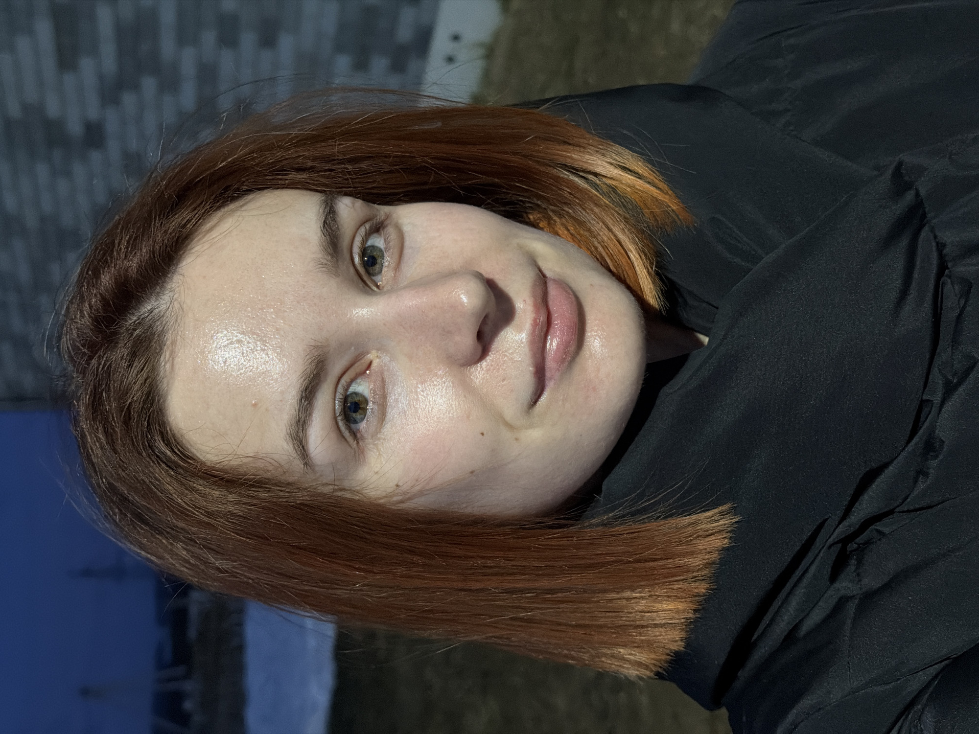 A close-up portrait of a young woman with fair skin and reddish-brown hair, possibly dyed. She has green eyes and is wearing a dark, possibly black, jacket. Her skin appears slightly shiny. The background includes a dark, possibly brick, wall and a blurry expanse of what appears to be dusk or a poorly lit area. Based on the query's information, the location is Chaikovsky, Russia.