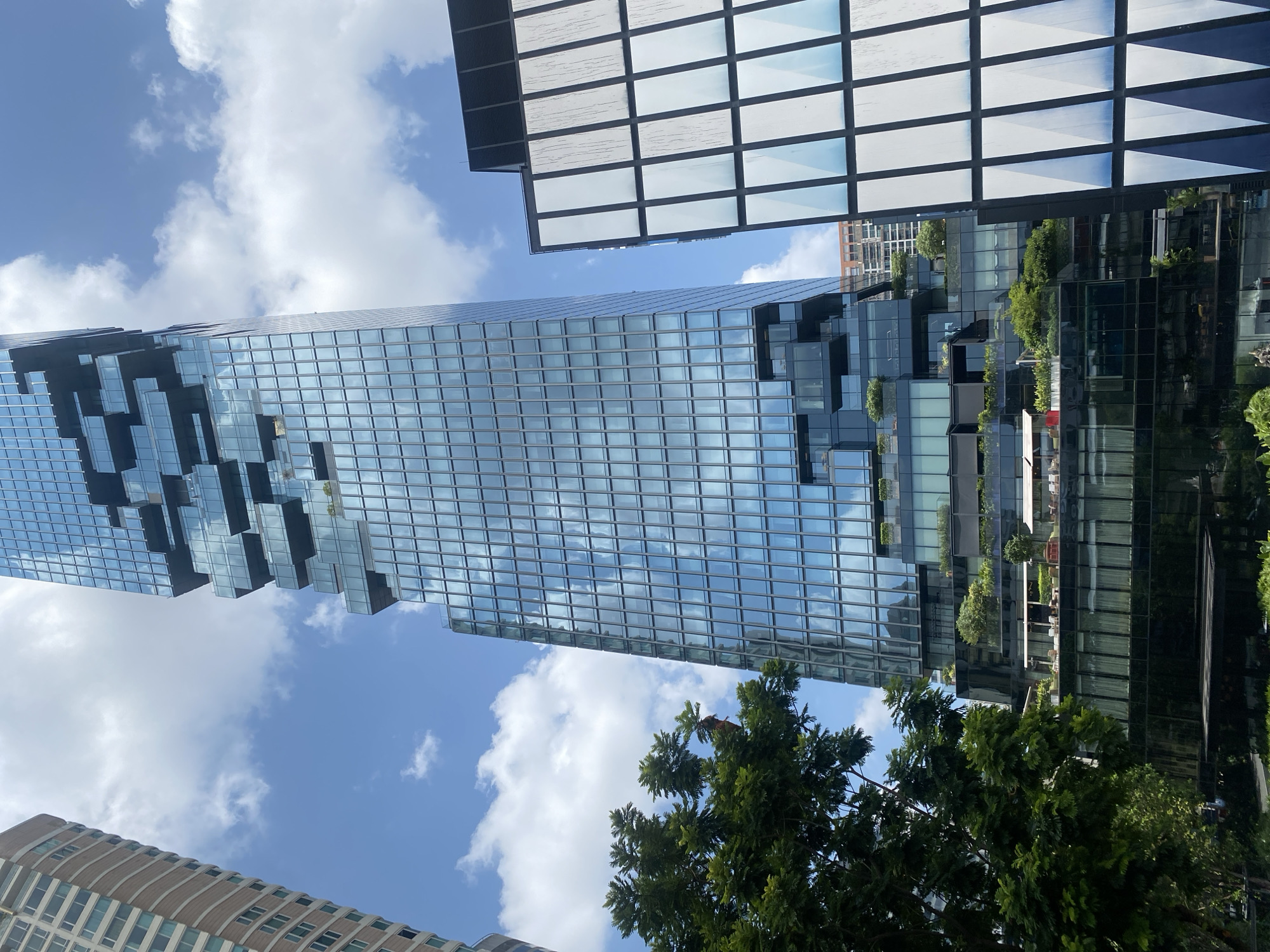 The image shows a view of the MahaNakhon building in Bangkok, Thailand, taken from a ground-level perspective. The skyscraper is composed of glass panels, some of which are cut away, giving it a pixelated appearance. The blue sky and white clouds are reflected in the windows. Part of another skyscraper, with a square grid facade, is visible in the upper right corner. Trees are present in the lower portion of the image. Part of another building can be seen in the lower left corner.