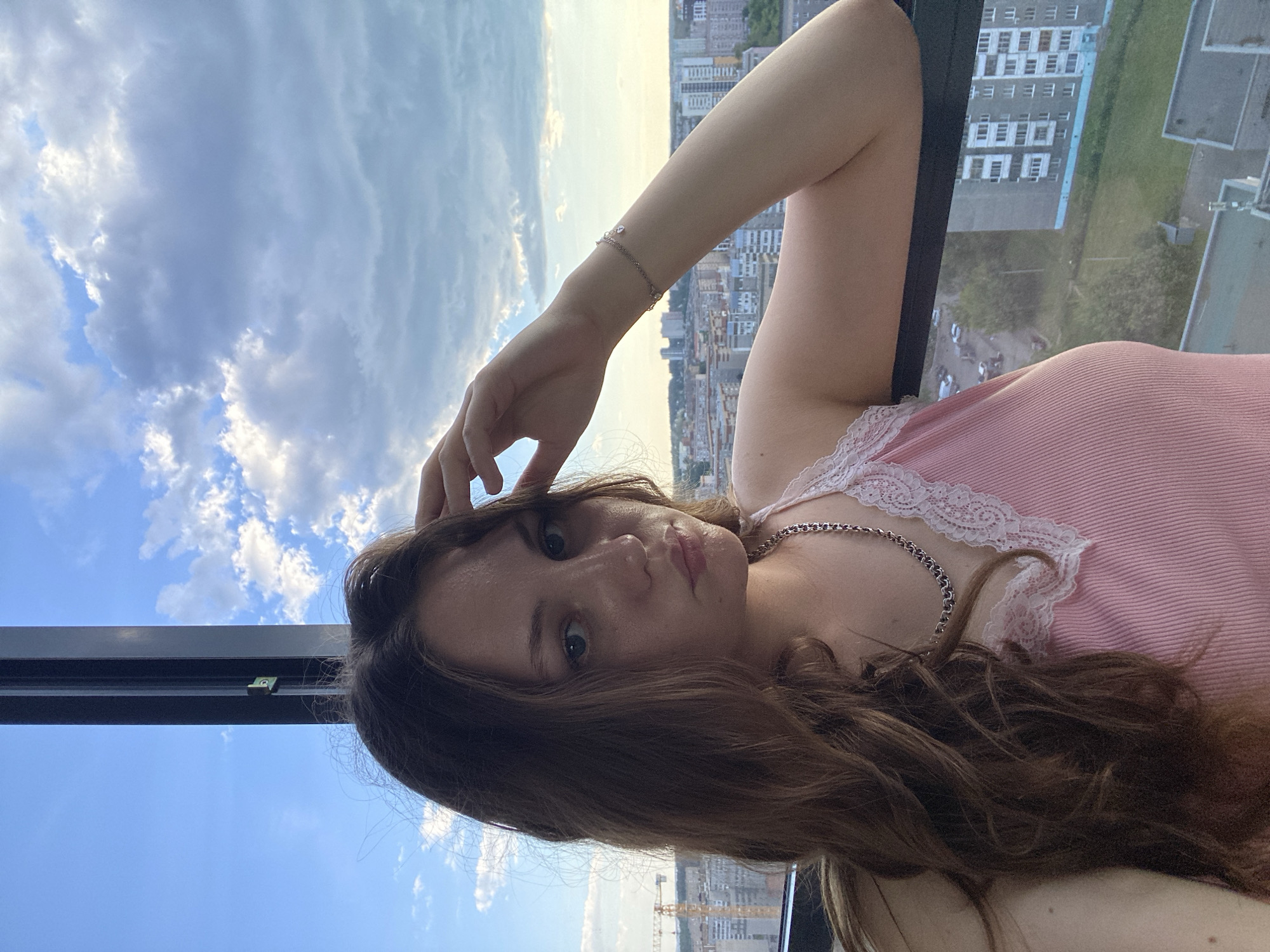 A young woman is pictured from the chest up, looking towards the camera with a neutral expression. She has long, wavy brown hair and is wearing a pink ribbed tank top with white lace trim. A silver chain necklace is visible around her neck. Her right arm is raised, with her hand resting on her head. She is wearing a bracelet on her right wrist. The background shows a cityscape under a cloudy sky. The presence of construction cranes suggests the image was taken in Izhevsk, Russia, a city known for its industrial and manufacturing sectors.