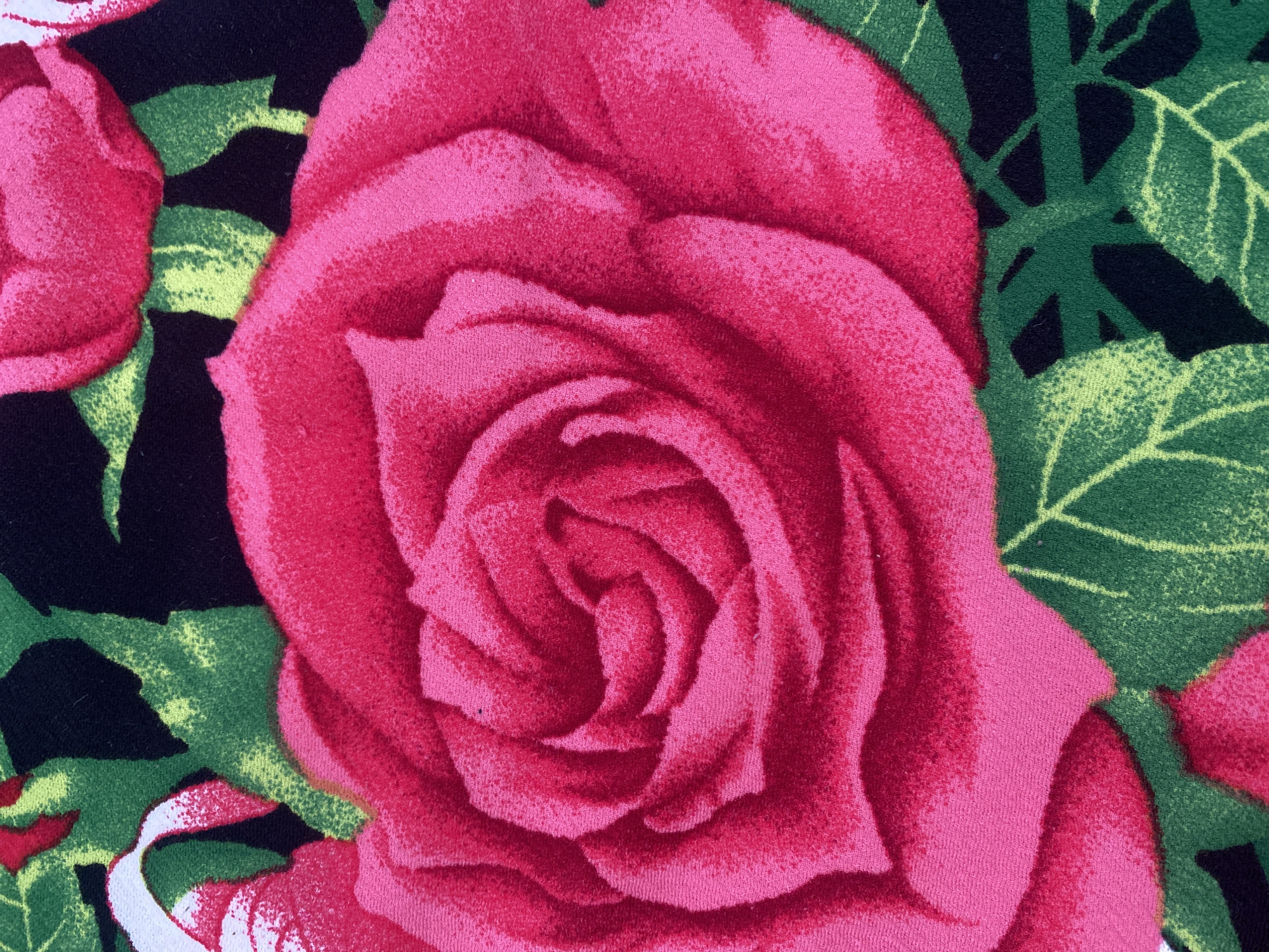 This close-up image displays a vibrant floral pattern on a textile fabric. The main subject is a large, stylized pink rose, dominating the center, with its layered petals rendered in varying shades of fuchsia and lighter pink, creating an impression of depth. Surrounding the central rose and in the background are other partial pink rose forms and numerous green leaves. The leaves exhibit a range of green hues, from dark forest green to bright lime green, and some show distinct vein patterns. The background of the pattern is a solid black, which provides a strong contrast to the bright pink and green elements. The fabric itself shows a discernible woven or knitted texture, suggesting a soft material. There are no people or specific activities depicted. The lighting is even, allowing the details of the print and fabric texture to be clearly seen. There is no visible text. The image captures a decorative pattern, potentially from clothing, upholstery, or a similar item, within the context of Davao City, Philippines.