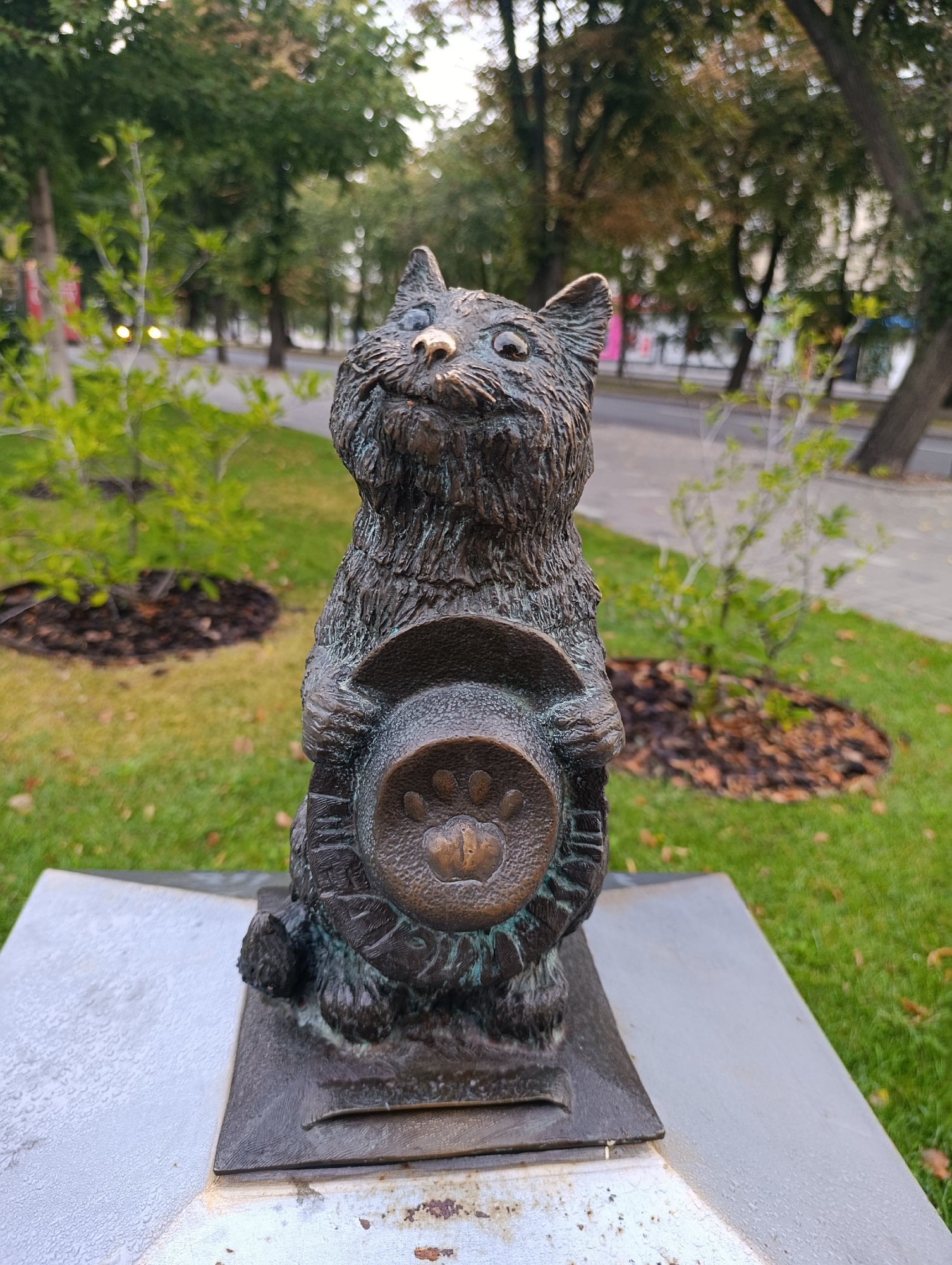 The image shows a bronze statue of a cat holding a hat with a paw print on it. The statue is situated outdoors on a paved platform surrounded by grass and trees, suggesting a park or public space. The cat statue is the central subject, depicted with a whimsical expression and textured fur. The hat it holds is round and appears to be a bowler hat. In the background, there are trees with green and autumnal foliage, and a road with some indistinct figures or vehicles. This statue is located in Cherkasy, Ukraine, and is known as the "Cat of Professor Sniezhnitsky."