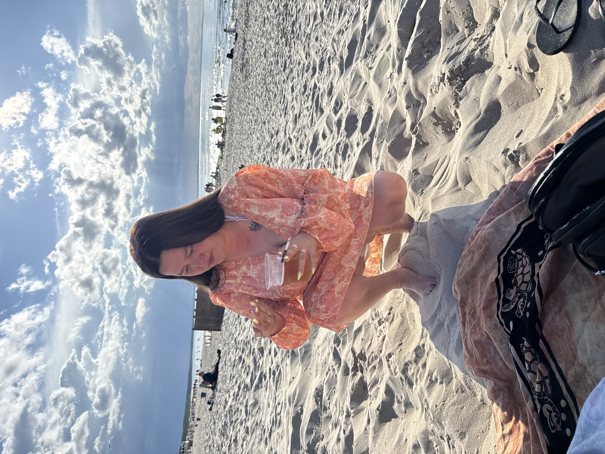 A woman is crouched on a sandy beach in Międzyzdroje, Poland, holding a clear plastic cup of amber-colored liquid in her right hand and what appears to be food in her left. She is wearing an orange floral-patterned dress over a white bikini top, with a tattoo visible on her left upper chest. Her fingernails are painted yellow. In front of her, on the sand, is a blanket with a black and orange pattern featuring sea turtles. A black purse and a pair of black flip-flops are also on the sand nearby. The beach is populated with other beachgoers in the background, some sitting and some standing, and the sea stretches to the horizon under a partly cloudy sky.