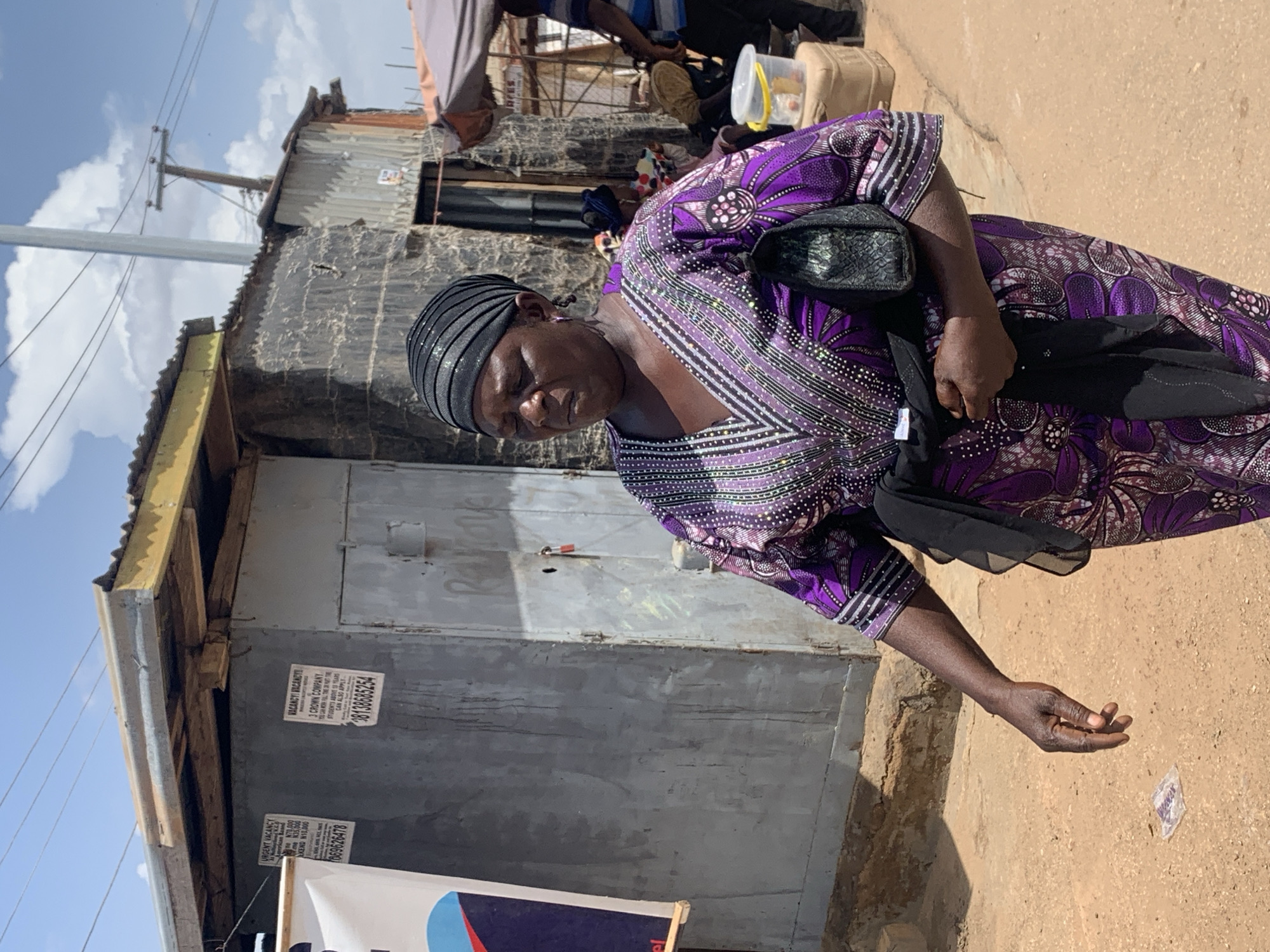 The image captures a scene in Gura Top, Nigeria, featuring a woman standing outdoors on what appears to be an unpaved street or pathway. She is the central subject, dressed in a vibrant purple patterned gown with embellishments and a black headwrap. She carries a black shoulder bag and extends her right hand, palm-up, towards the dusty ground, suggesting she is soliciting or receiving something. Her facial expression appears serious or intent.
In the background, there are simple, weathered structures, likely residences or small businesses, constructed from corrugated metal sheets and concrete blocks. Exposed timber supports are visible on the buildings. The sky above is bright with scattered white clouds, indicating it is daytime with strong sunlight casting distinct shadows. Power lines and a utility pole are visible against the sky.
Affixed to the wall of one building are two hand-posted advertisements. One poster reads "URGENT VACANCY," specifying amounts like "N70.000" and "N10,000," along with a phone number "08069626478." The second poster declares "VACANCY! VACANCY! 3 CROWN COMPANY," stating that "STUDENTS ABOVE 10 YEARS CAN ALSO APPLY," and includes the phone number "08138035254." A partial word, "Rahove," is also discernible. A small, crumpled object rests on the ground near the woman's hand. Other indistinct people are faintly visible further in the background. The image is rotated 90 degrees clockwise.