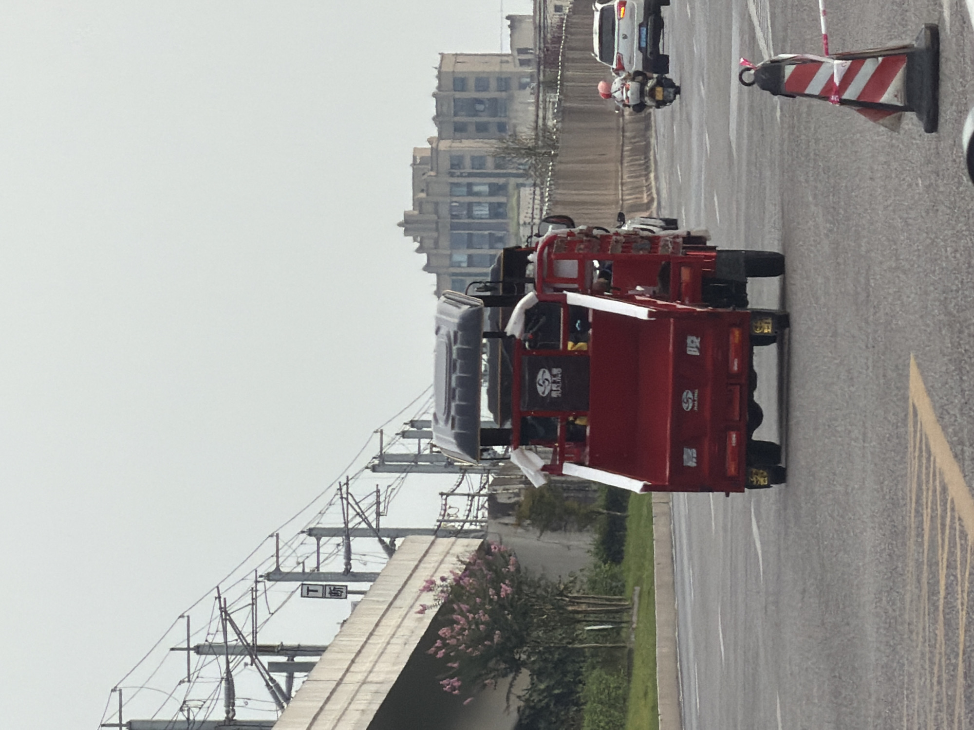 A red three-wheeled vehicle is traveling on a multi-lane road in Jiangjin, China. The vehicle has a covered cab and an open cargo area. A white SUV and a motorcycle with a person wearing a pink helmet are also on the road. In the background, there are buildings and overhead railway tracks. The weather appears overcast.