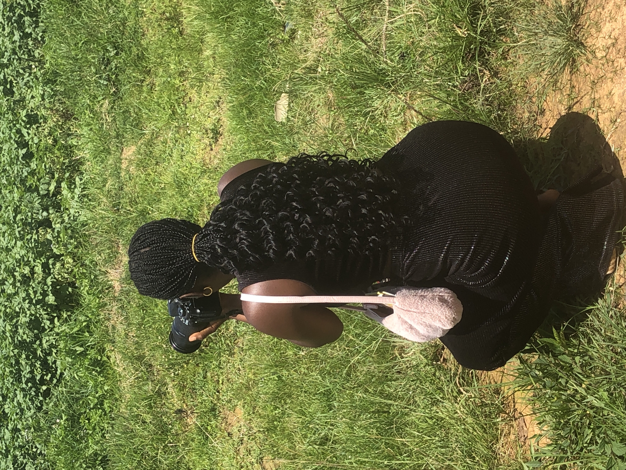 In Gwafan, Nigeria, a woman is photographed from behind, crouching in a field of green grass and foliage. She is wearing a black, ribbed dress and has her dark hair in braids with a golden tie. A fuzzy pink purse hangs from her shoulder, and she holds a black camera to her eye, appearing to be taking a photo of something in front of her. The ground around her is a mix of dirt and grass.