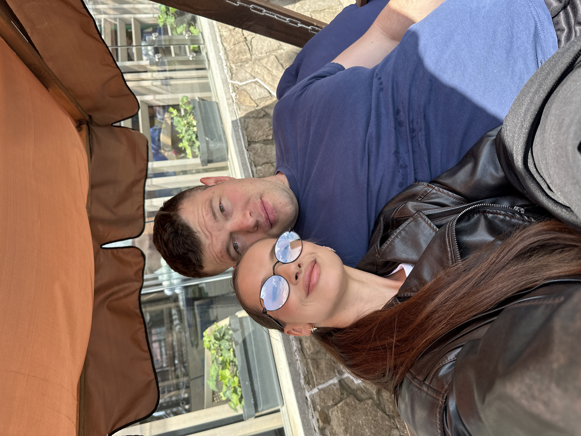 A man and a woman are taking a selfie on a swing set under an orange awning. The woman is wearing a black leather jacket and round sunglasses, her long brown hair is pulled back in a ponytail. The man is wearing a dark blue t-shirt. They are seated close together, and both are looking at the camera. In the background, there is a stone wall and a window with planters containing greenery. The scene appears to be outdoors, possibly at a cafe or restaurant with outdoor seating.