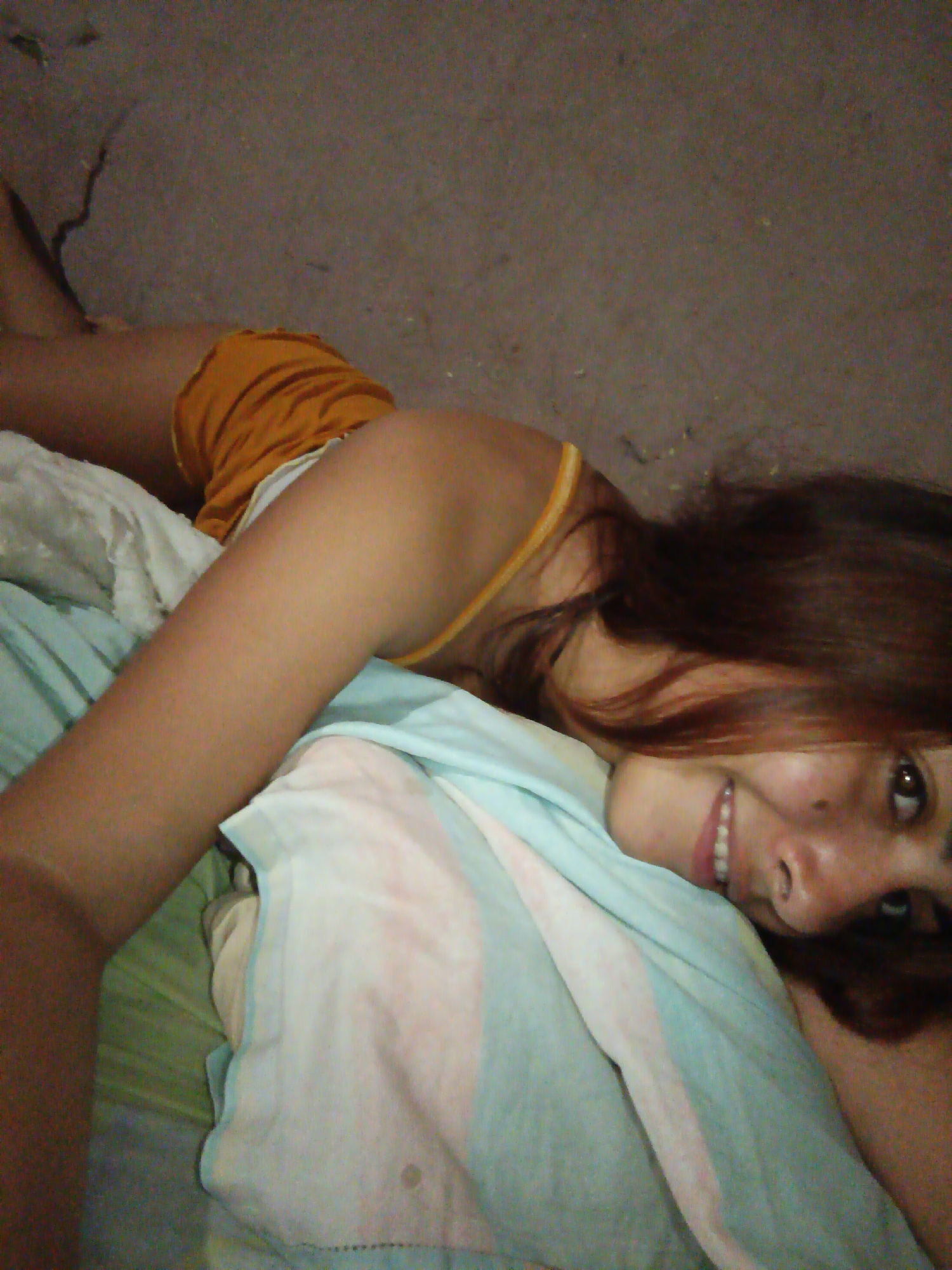 The image is a close-up, low-angle selfie of a young woman lying down. She has medium-toned skin, dark brown hair, and is wearing an orange top. She is lying on a bed with light blue and white striped bedding, and a green blanket underneath. Her head is tilted, and she is looking at the camera with a smile. The background appears to be a textured, dark surface, possibly a wall or floor. The image was taken in Juan Griego, Venezuela.