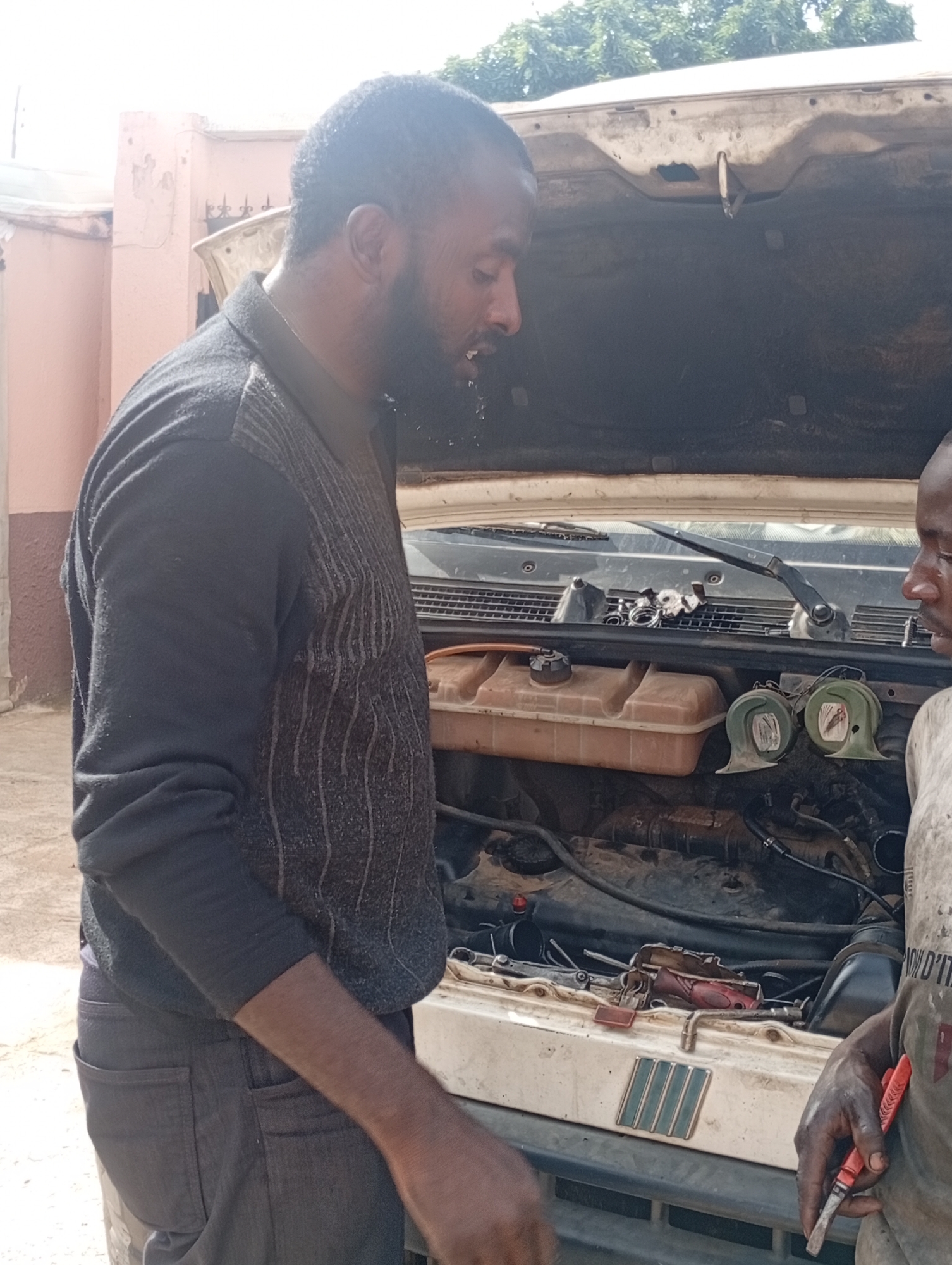 Two men are working on the engine of a car with the hood open. The man on the left, with a beard and wearing a dark sweater, is looking down into the engine. The man on the right, wearing a light-colored t-shirt, is holding a red tool with a silver tip, possibly a screwdriver or chisel, and is also looking at the engine. The engine compartment is dirty and appears to be a mechanical repair in progress.
The scene appears to be outdoors, possibly a residential area or a mechanic's workshop, given the presence of a building in the background with pink walls and a fence. Trees are visible above the car. The lighting suggests it is daytime, with bright sunlight. The overall atmosphere seems focused and practical, indicative of a repair task. There is no visible text in the image. The location context is Jos, Nigeria.