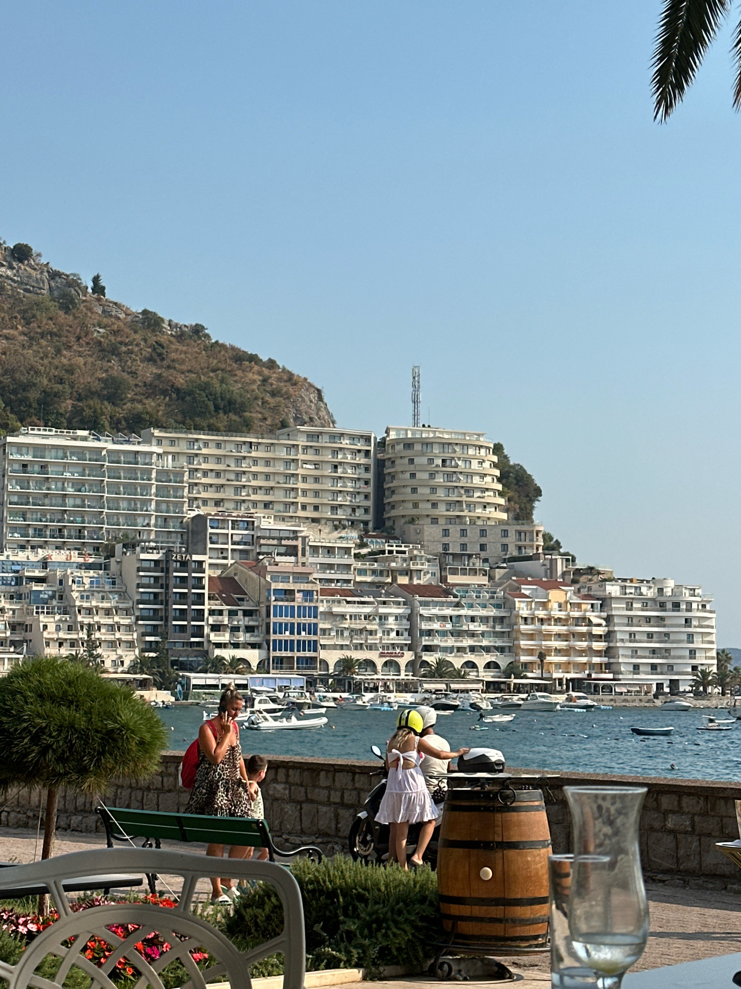 In Bečići, Montenegro, a sun-drenched scene unfolds along the waterfront, dominated by a sprawling hillside adorned with numerous white buildings. These structures, ranging from hotels to apartment complexes, cascade down the slope towards the azure sea, where a flotilla of small boats is anchored. In the foreground, a paved walkway lines the water's edge, separated from the promenade by a low stone wall.
On this walkway, two individuals, a woman and a child, are engaged in conversation. The woman, wearing a leopard-print dress and a red backpack, is talking on her phone. Beside her, a young child in a light-colored dress stands next to a parked scooter. The child is wearing a yellow helmet. A wooden barrel and a glass of water are positioned on a nearby table, hinting at a relaxed outdoor dining or seating area. To the left, a lush green tree provides a touch of nature, while a park bench offers a place to rest. The sky above is a clear, cloudless blue, completing this idyllic coastal vista.