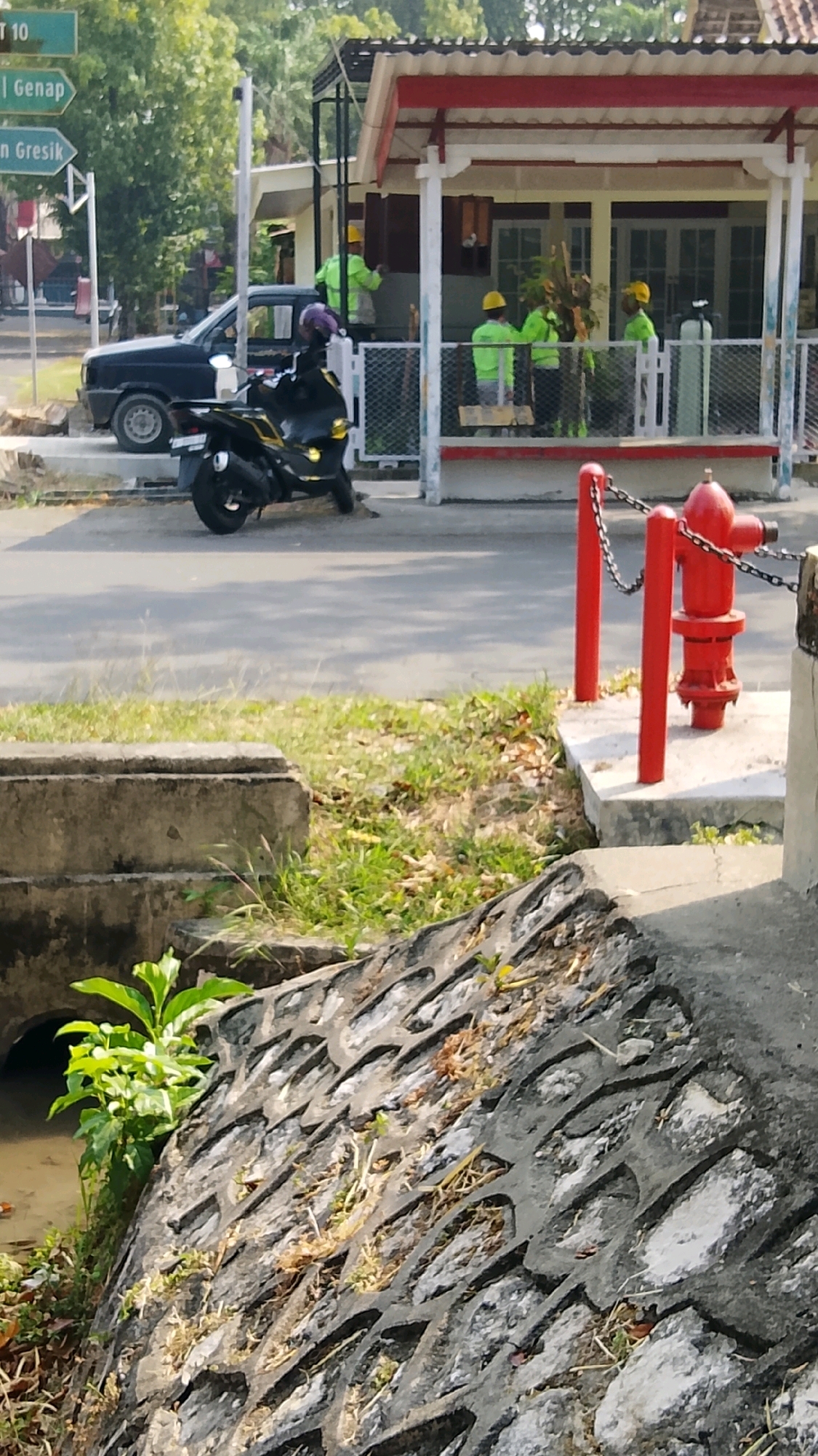 In Tlogobendung, Indonesia, a street scene unfolds under a bright sky. On the left, a black pickup truck is parked near a signpost indicating directions to "Lamongan" and "Gresik." Adjacent to the truck, a black scooter with a purple helmet on its seat is positioned on the asphalt. In the background, a building with a white facade and a red-trimmed roof is partially visible. Several individuals, wearing bright green shirts and yellow hard hats, are seen working around this building, some appearing to be engaged in outdoor tasks. A red fire hydrant with a chain fence stands prominently in the foreground, situated on a concrete base. The area is bordered by grassy patches and what appears to be a drainage ditch with a concrete structure, where a small green plant grows.