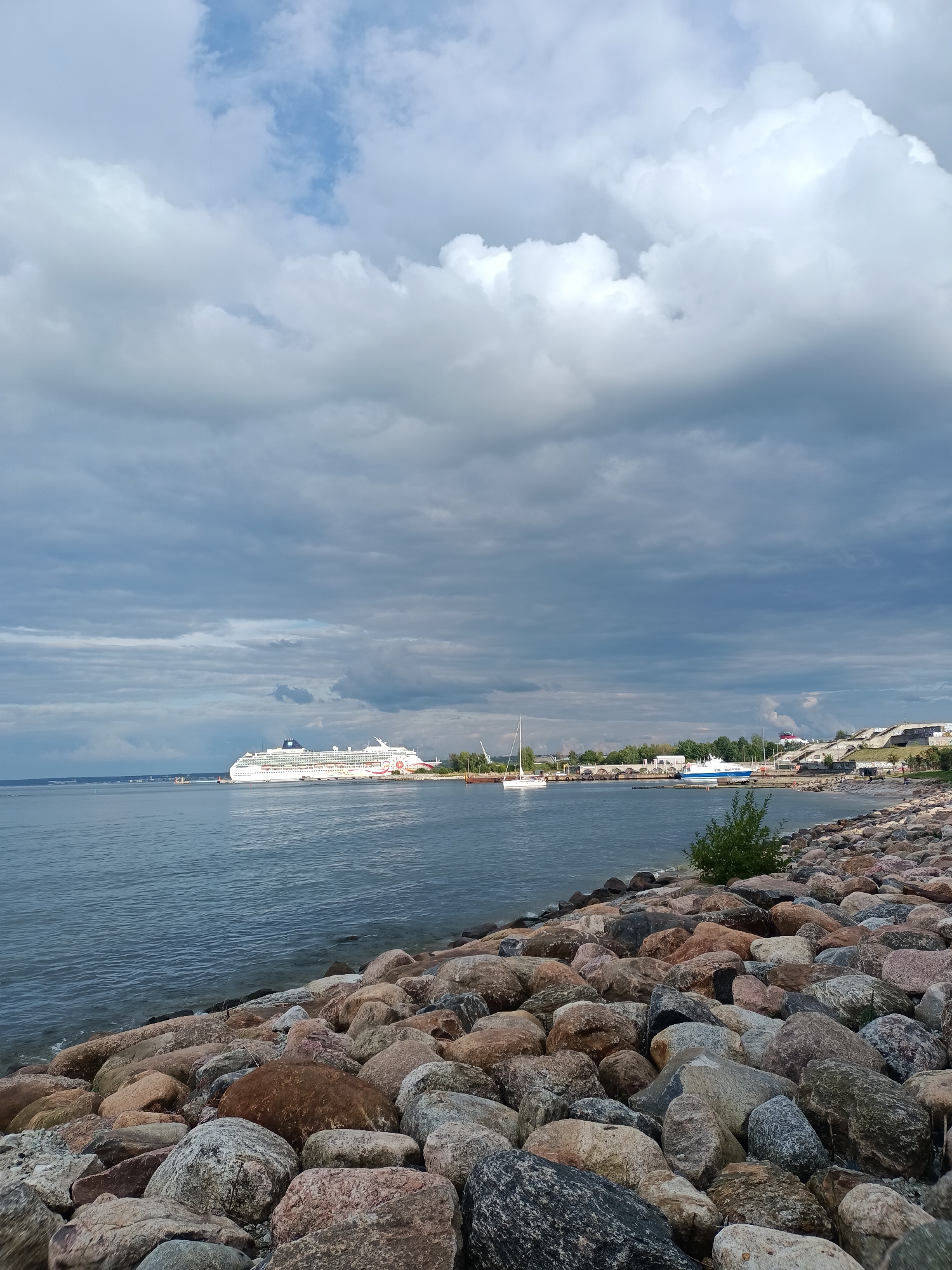 The image shows a view of the Tallinn Bay in Tallinn, Estonia, from a rocky shoreline. In the middle ground, a large white cruise ship with colorful accents on its stern is docked or anchored. Several other boats, including a sailboat, are visible in the harbor area. The shoreline is covered with numerous large rocks and boulders. In the background, there are buildings, trees, and a tiered staircase structure on a hill. The sky is filled with dramatic, cloudy formations, with patches of blue sky visible. The water is calm and reflects the sky and the surrounding scenery. There are no people clearly visible in the image, but some faint figures can be discerned on the distant shore near the staircase. The overall impression is a scenic view of a coastal city with maritime activity under a cloudy sky.