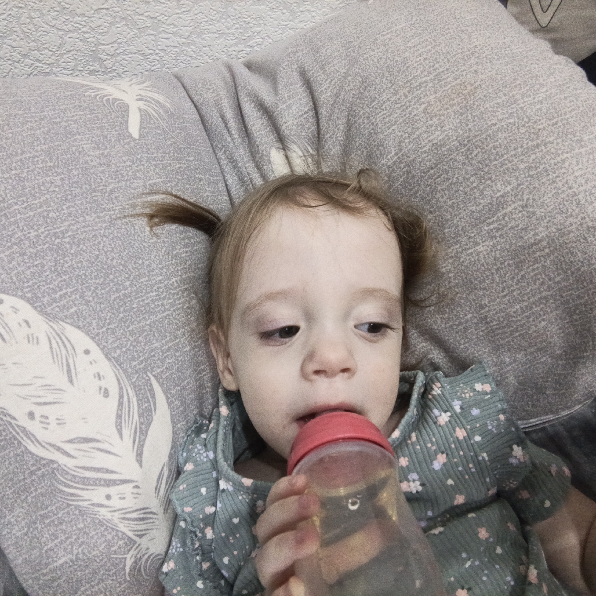 An overhead close-up shot features a young child, likely a toddler, reclining indoors, possibly on a bed or couch. The child has light brown hair styled in two small pigtails and is wearing a greenish-blue ribbed top adorned with small floral patterns in pink, purple, and white.
The primary activity is the child drinking from a translucent plastic sippy cup or bottle with a red lid, which contains a light brown or yellow liquid. The child's eyes are cast downwards and slightly to the side, indicating focus on the drink, with a neutral and calm expression.
Behind the child are two grey pillows, one prominently featuring a large white feather design. The background includes a textured light grey or off-white wall or wallpaper. The overall setting appears to be a comfortable, domestic environment. While the provided location context is Марковский, Russia, the image itself does not contain specific visual cues to denote this geographic location. No text is visible in the image.