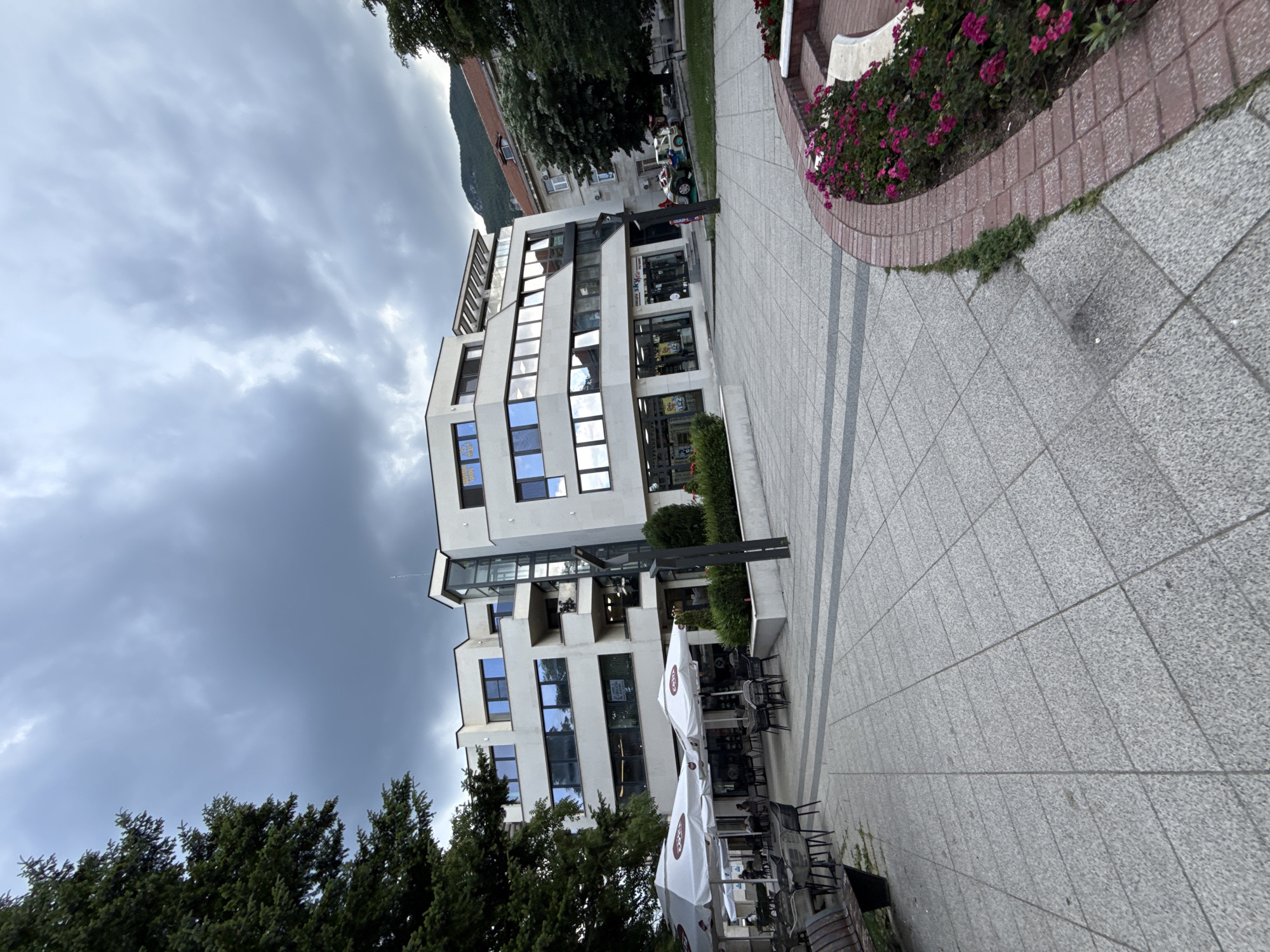 An exterior shot depicts a modern, multi-story white building with extensive glass windows in Vratsa, Bulgaria, under a cloudy sky. In the foreground, a paved plaza features outdoor seating with umbrellas, tables, and chairs, suggesting a cafe or restaurant. To the left, a large evergreen tree stands, and to the right, a flower bed with vibrant pink flowers borders the plaza. The building's facade is a mix of white panels and reflective windows, with balconies on upper levels. Behind the modern building, an older building with a red-tiled roof is visible. The overall scene conveys a public urban space with commercial activity.